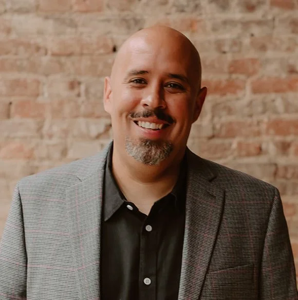 A man with a bald head and goatee smiling, wearing a gray plaid blazer and black shirt against a brick wall background.