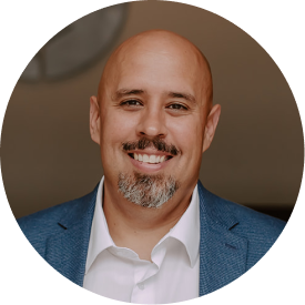 A man with a bald head and goatee smiling, wearing a gray plaid blazer and black shirt against a brick wall background.