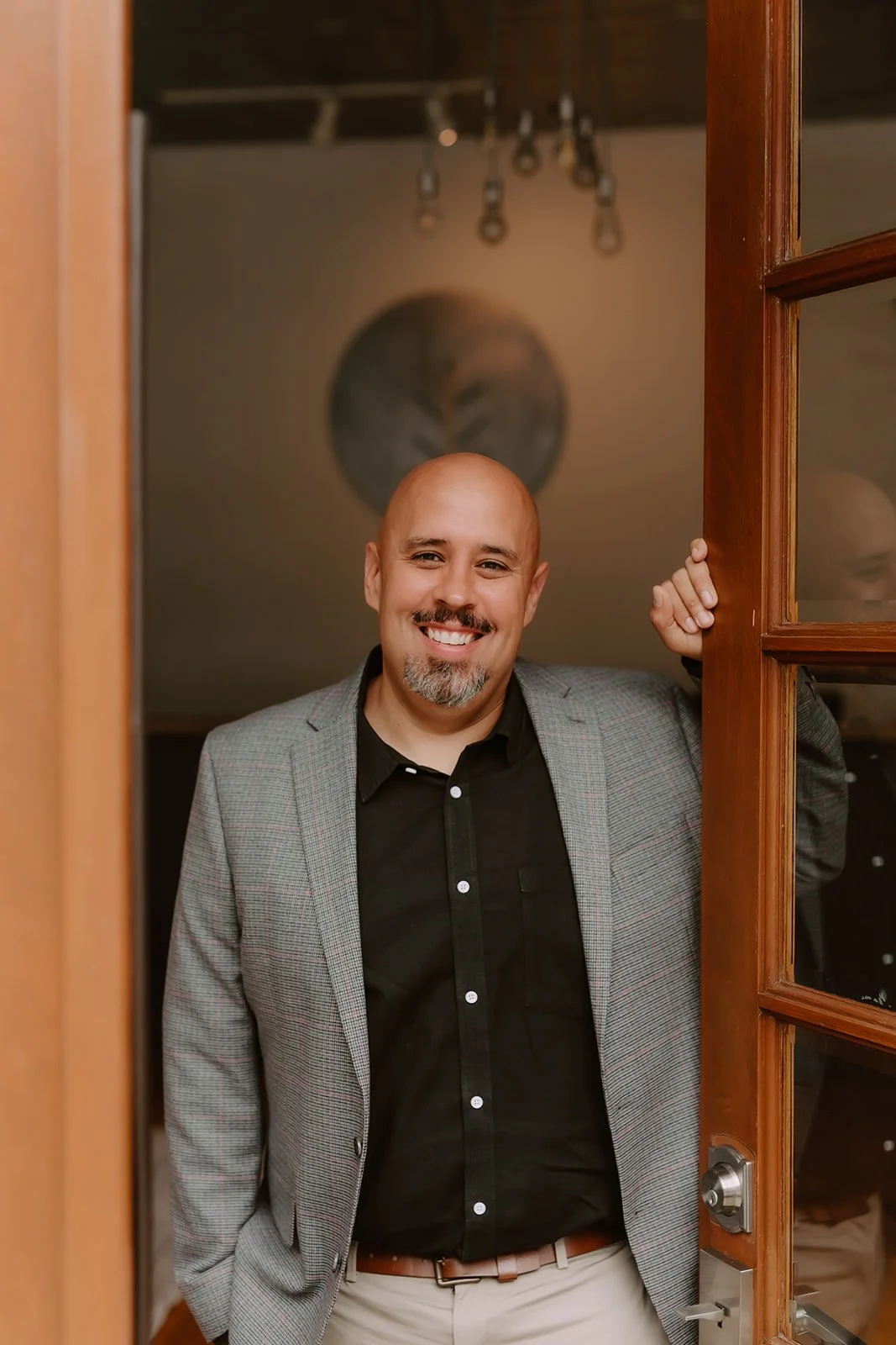 A man with a shaved head, smiling, stands behind an open wooden door, wearing a gray blazer over a black shirt and beige pants, in a warmly lit room.
