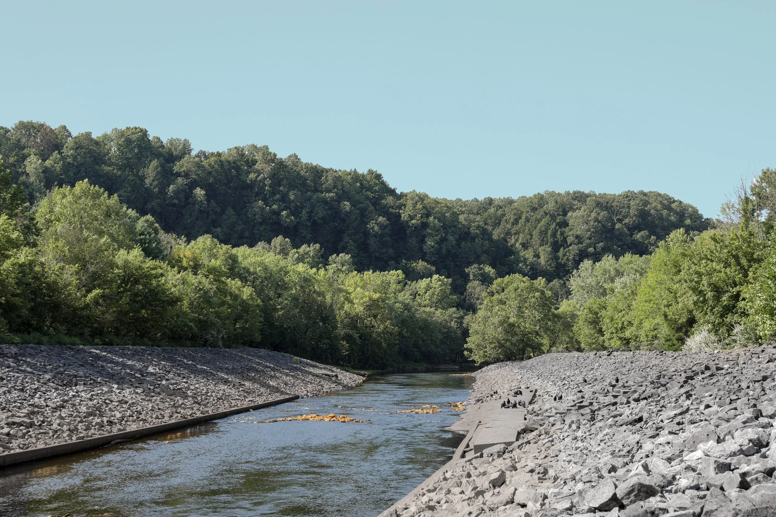 Stream running through a rocky riverbank with green trees and hills in the background under a clear blue sky.