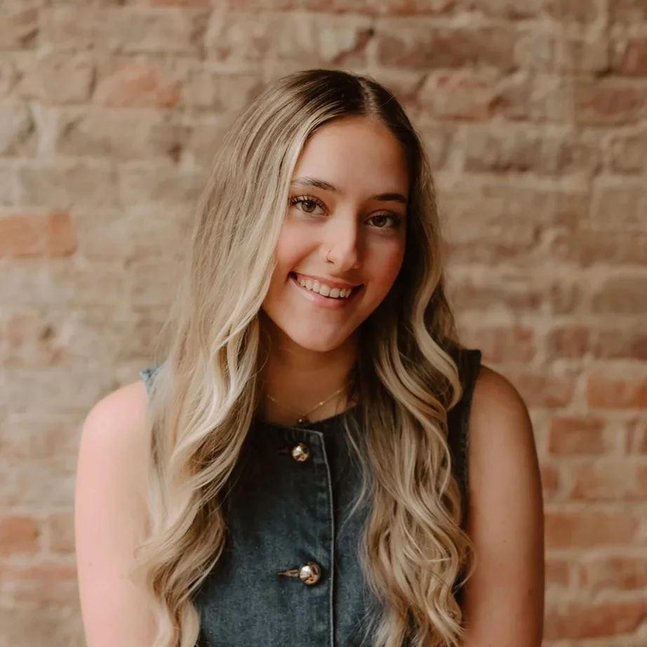 A young woman with long, wavy blonde hair and light skin, smiling at the camera, standing against a brick wall background.