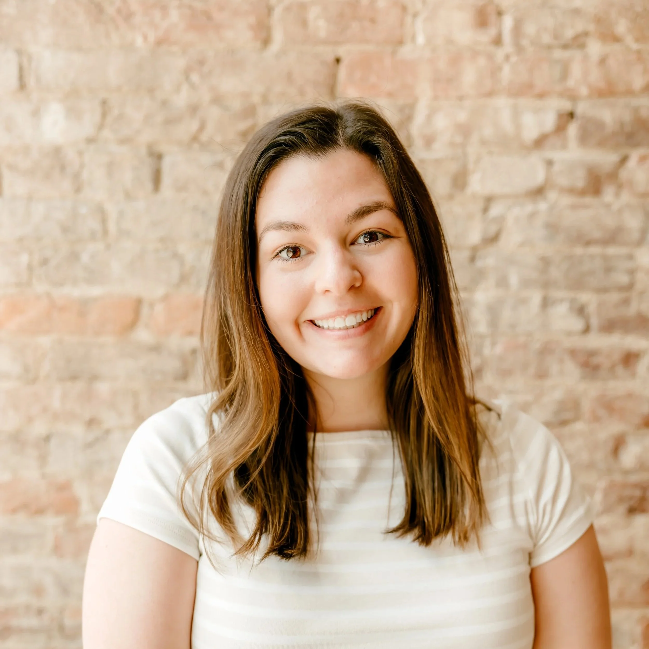 A woman with wavy brown hair wearing a black Adidas sports jacket with a Sharlow & Associates logo, smiling in front of a brick wall.