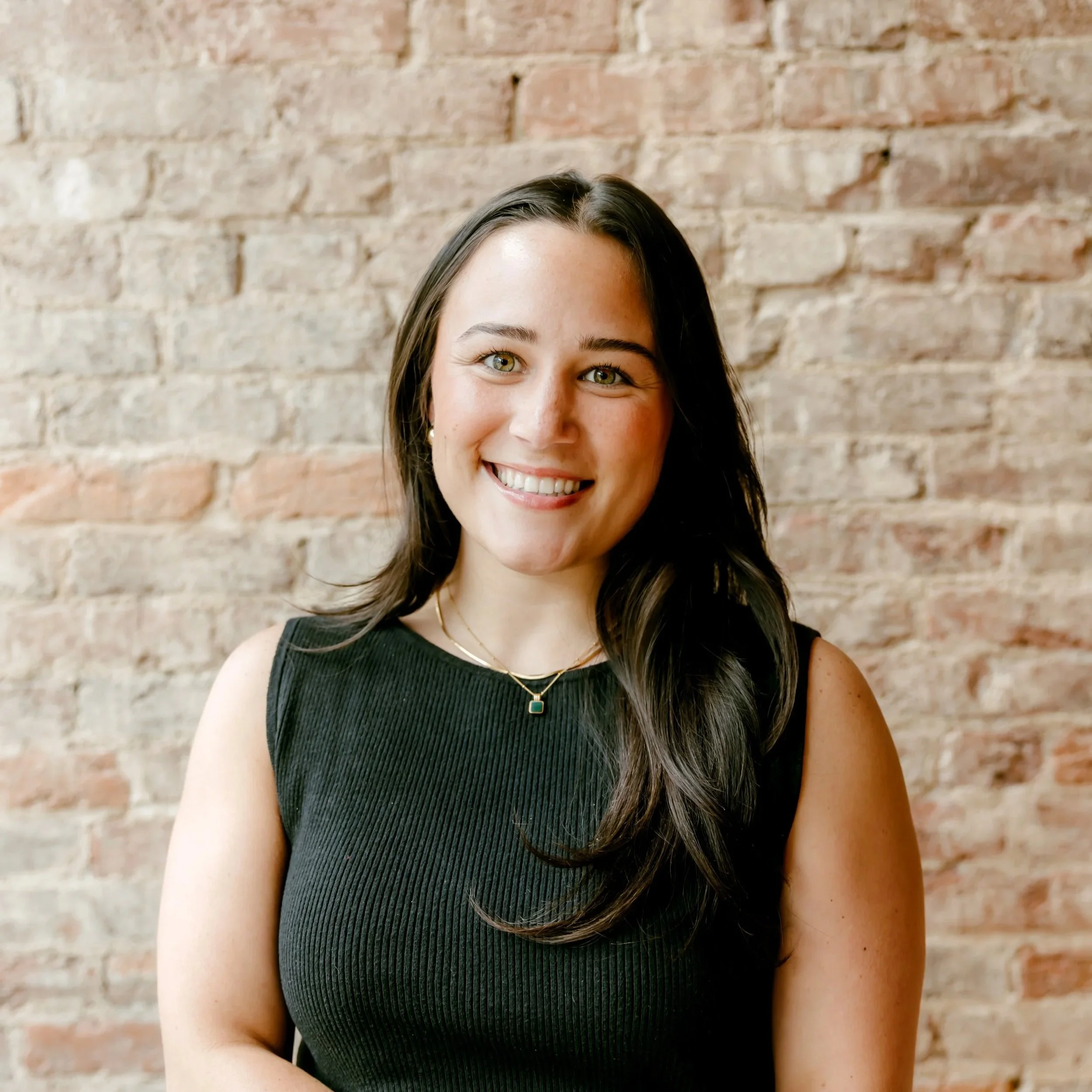 A young woman with long dark hair and a bright smile, wearing a gray t-shirt, standing in front of a brick wall.