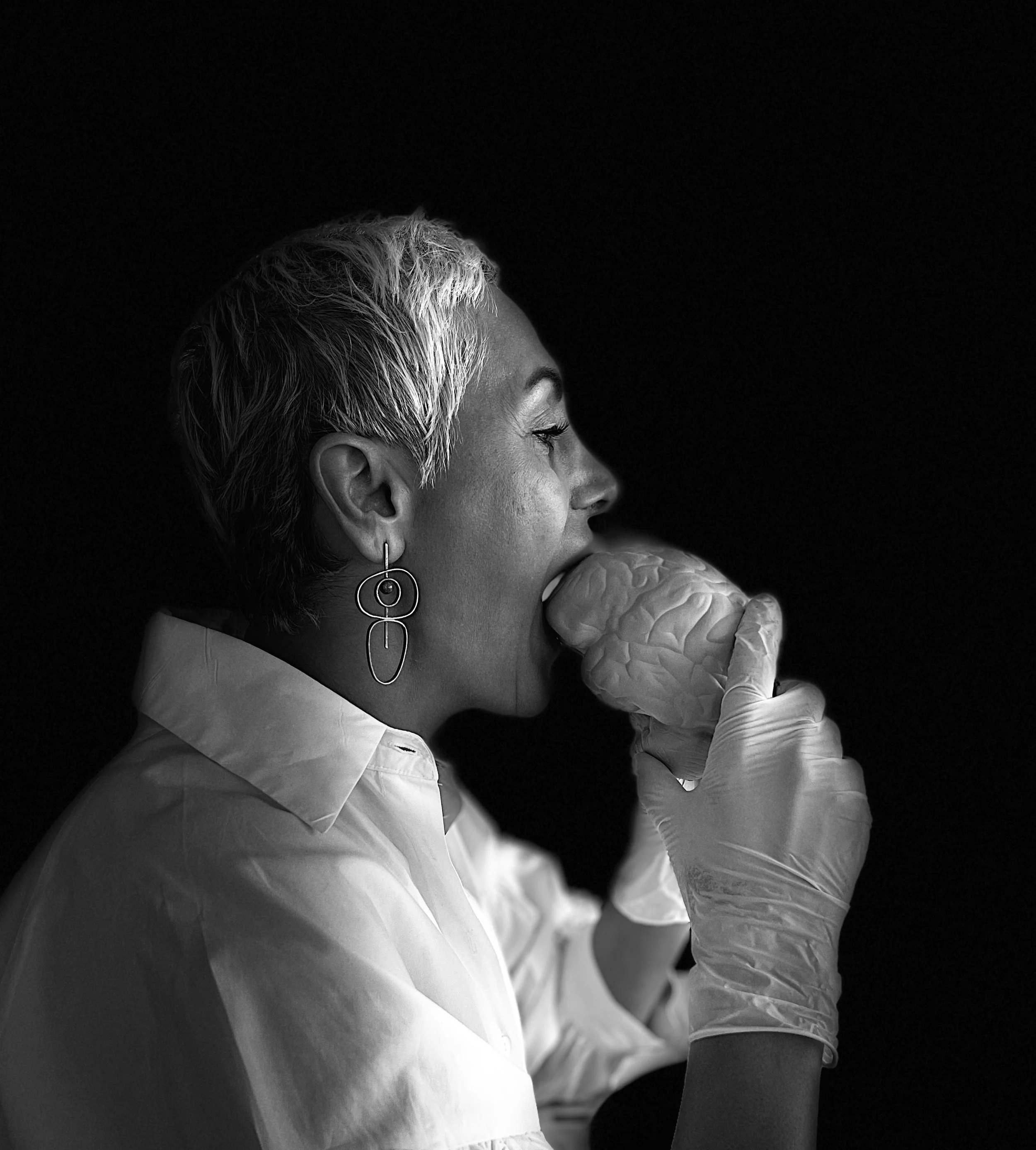 Black and white photo of a woman with short, platinum hair, wearing a white shirt and hoop earrings, drinking from a brain-shaped object.