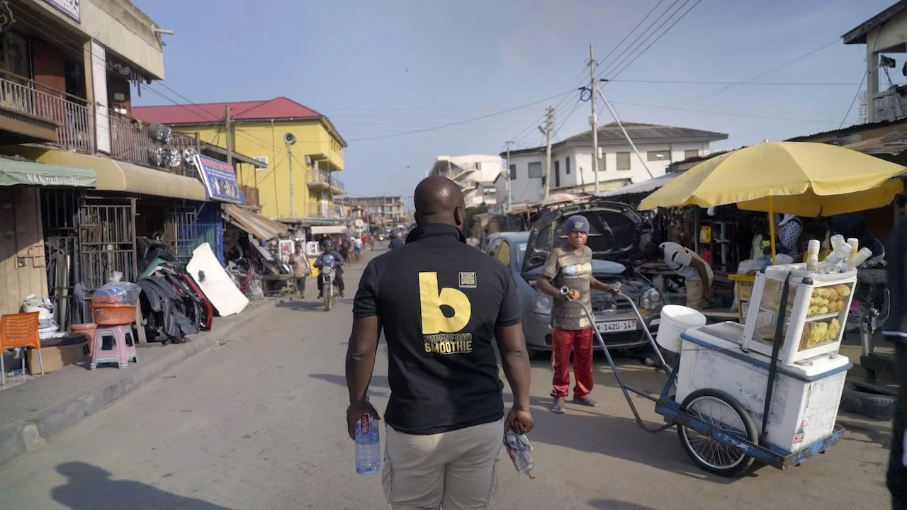 A busy street scene with shops, vendors, and pedestrians. The man in the foreground is walking away, holding a water bottle and a cloth. Other people are selling items under a yellow umbrella, and there is a food cart nearby. Buildings are on both si