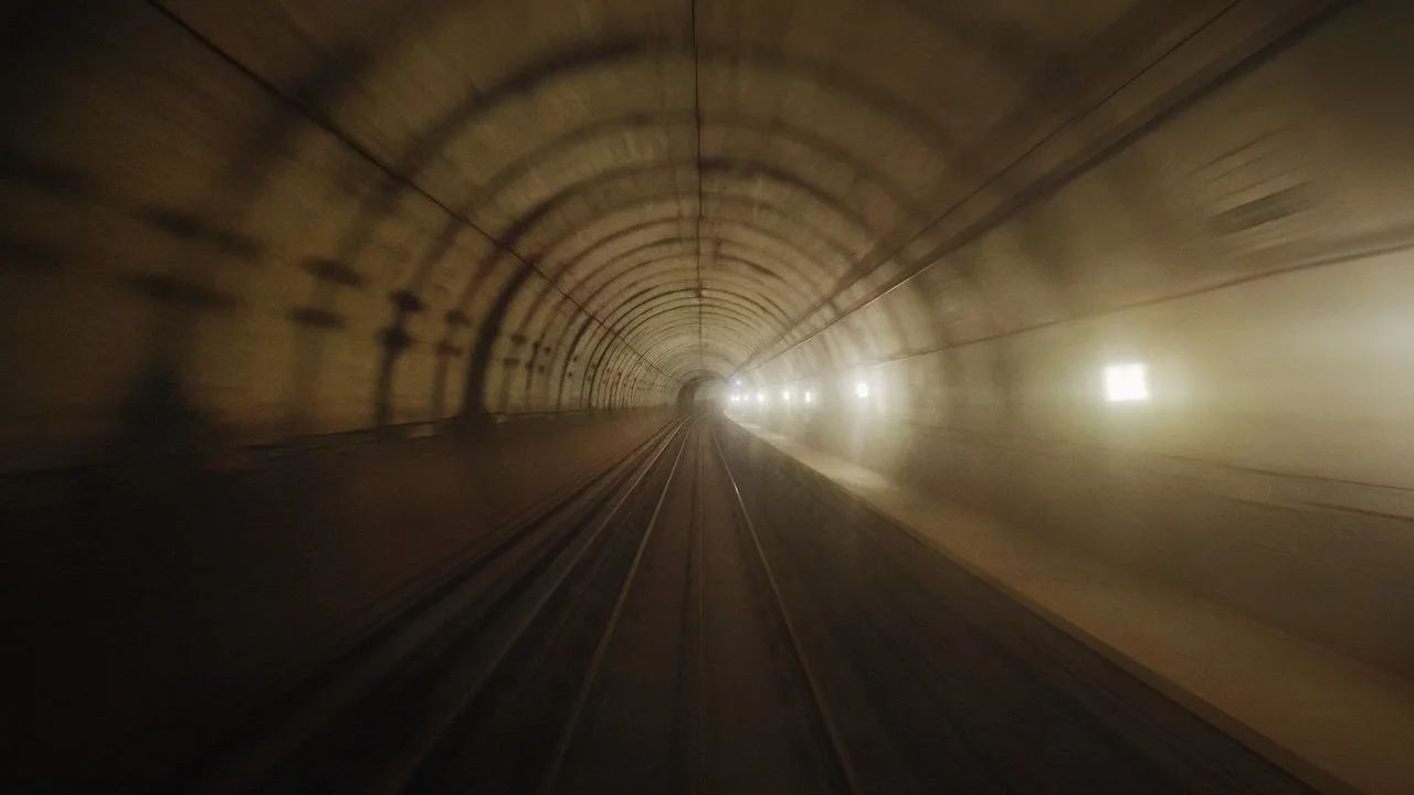 View of a tunnel with train tracks running through it, illuminated by wall-mounted lights, with a bright light at the end of the tunnel.