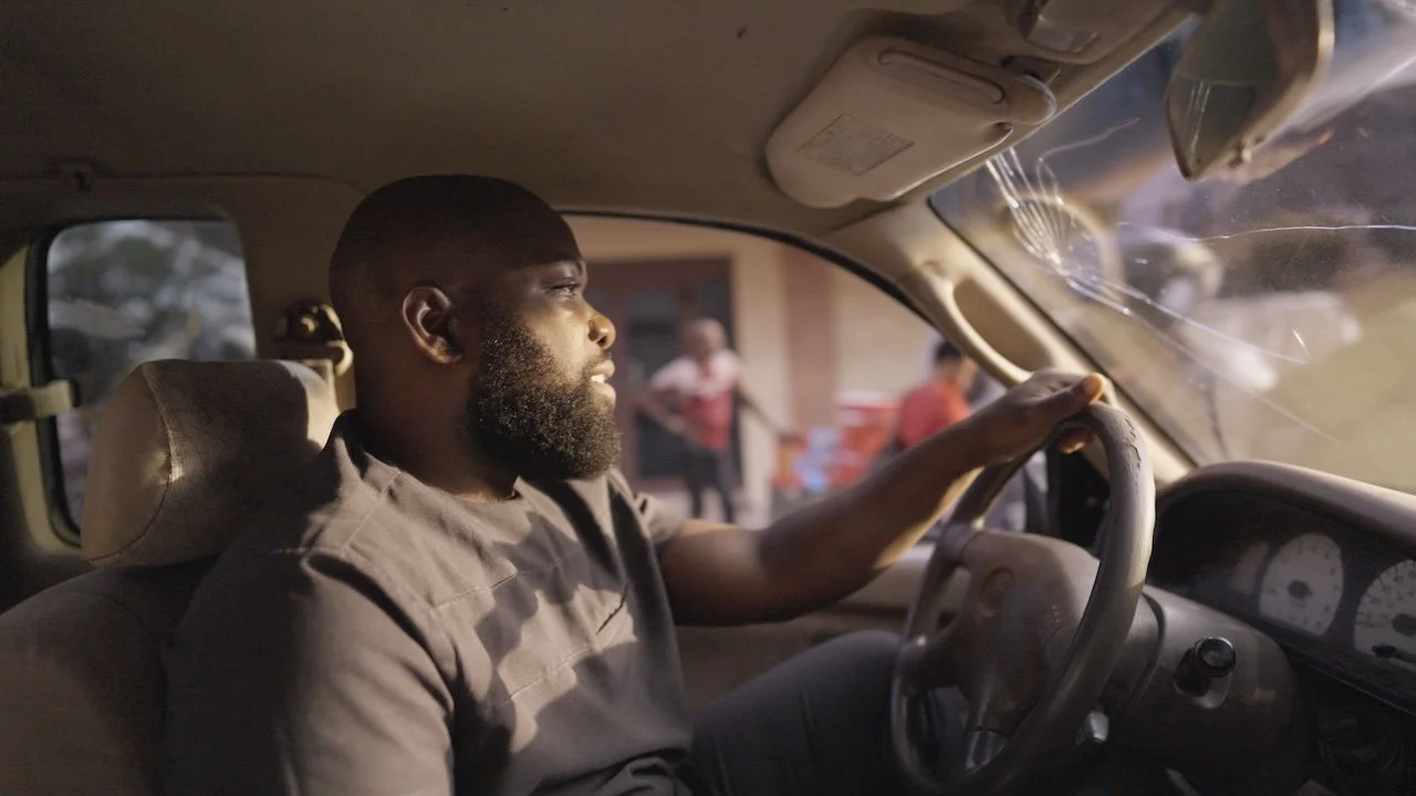 Man with beard driving a vehicle, looking focused, with people standing outside in the background.