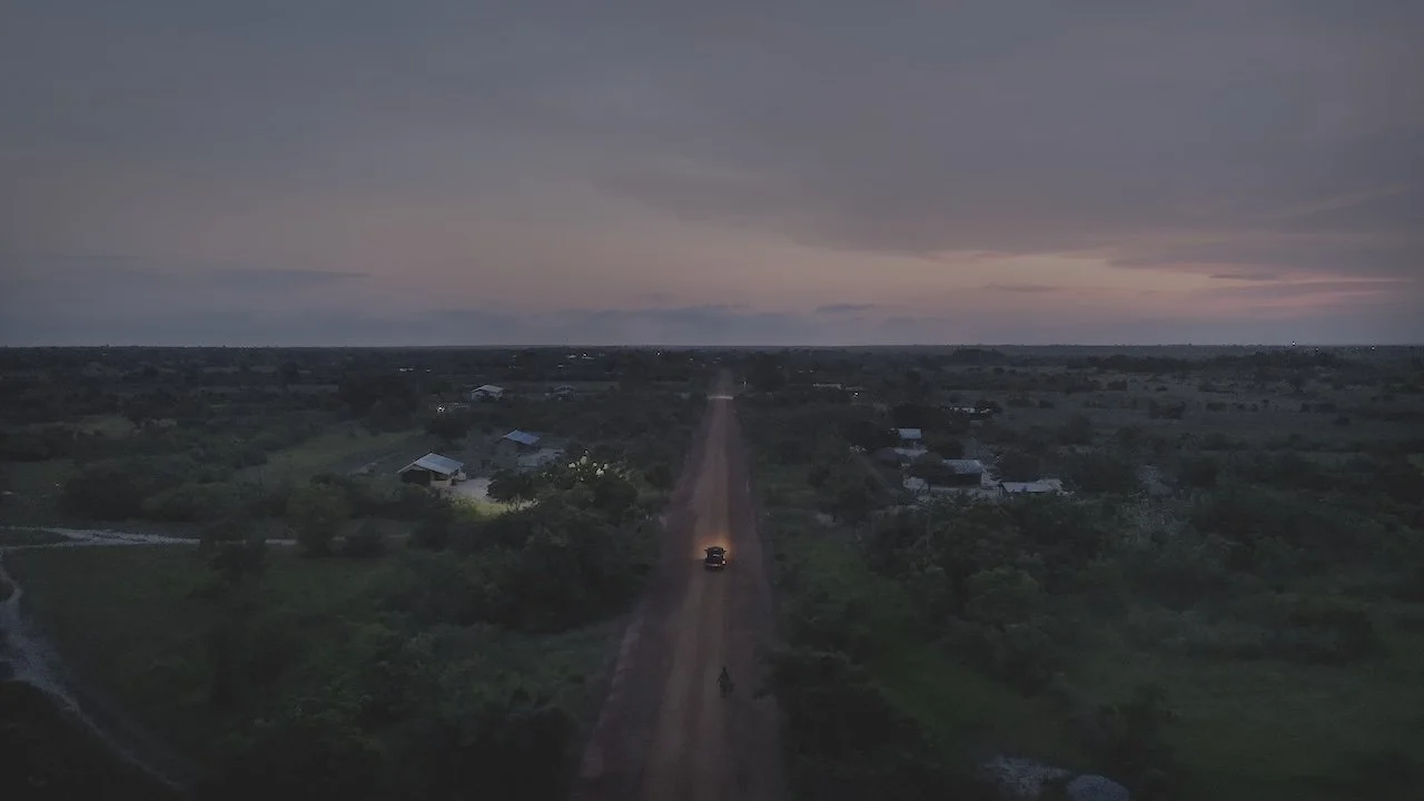 Dirt road stretching into the distance during sunset, with a vehicle and motorcycle on the road and small buildings on the sides, under a colorful sky.