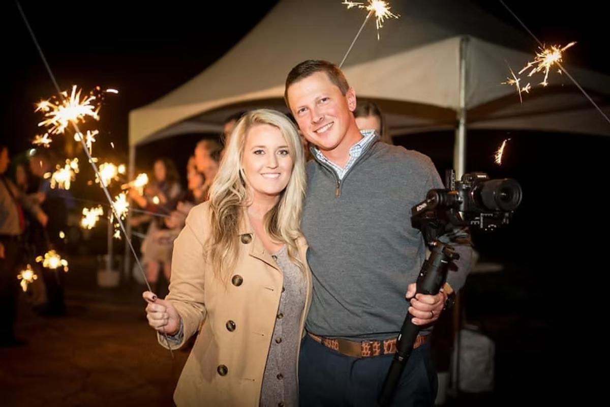 A smiling couple celebrating at night with sparklers, with a crowd and a white tent in the background.