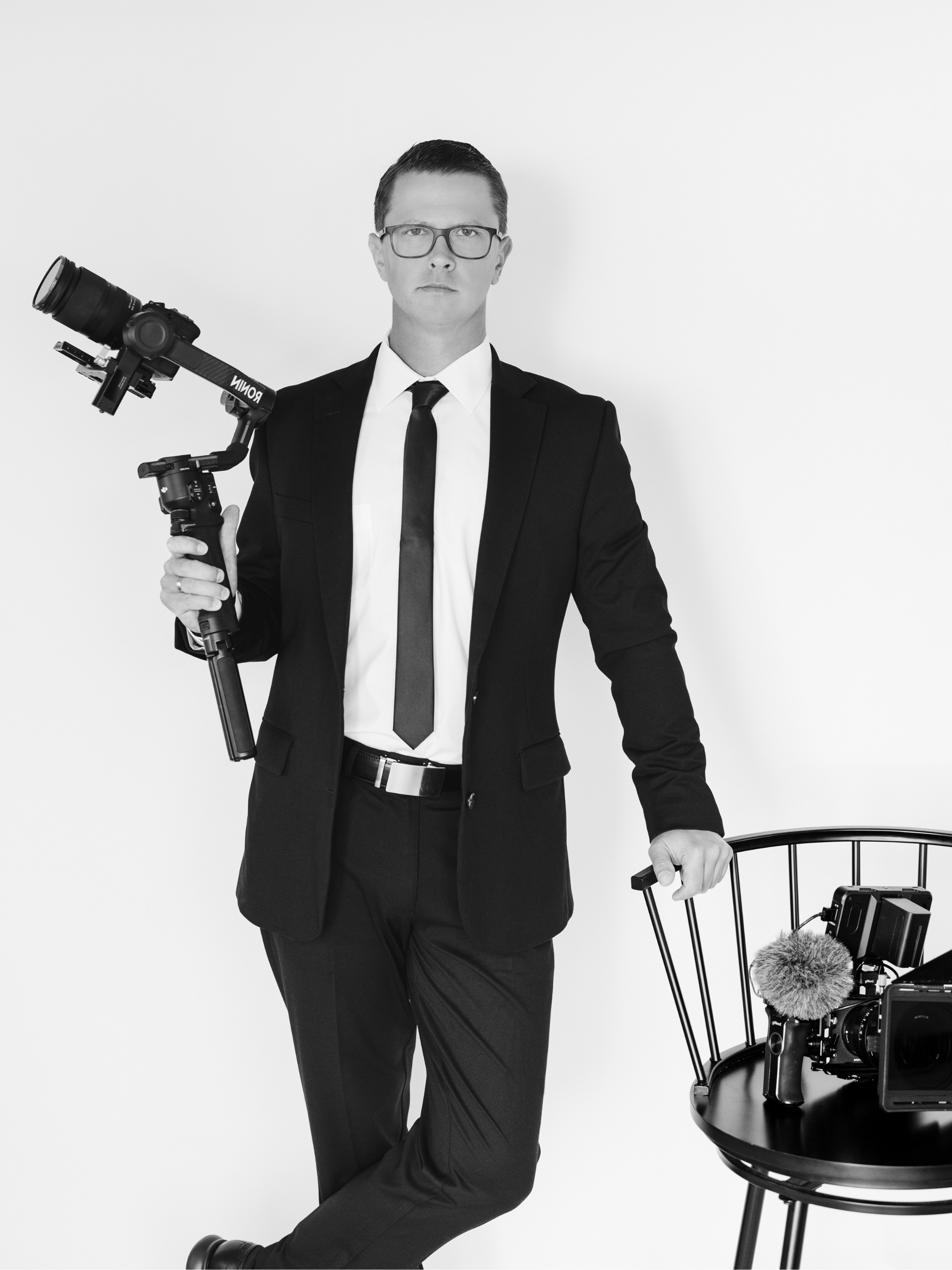 A man in a black suit, white shirt, and black tie holding a camera with a stabilizer. There is a chair with additional camera equipment and a microphone nearby.