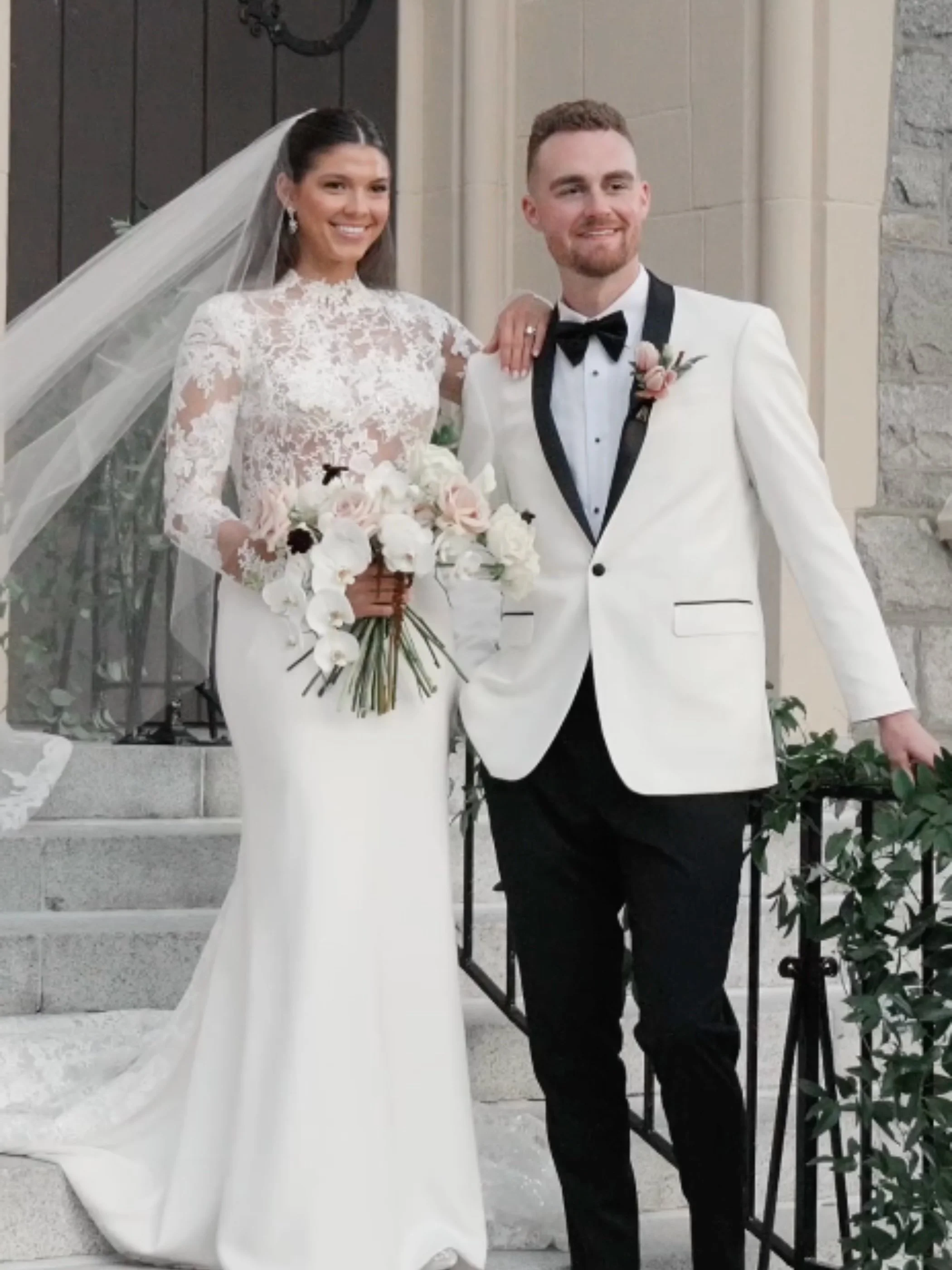 Bride and groom standing on church stairs, smiling, with the bride holding a bouquet of flowers, bride in a lace wedding gown and veil, groom in a white tuxedo jacket with black pants and bow tie.