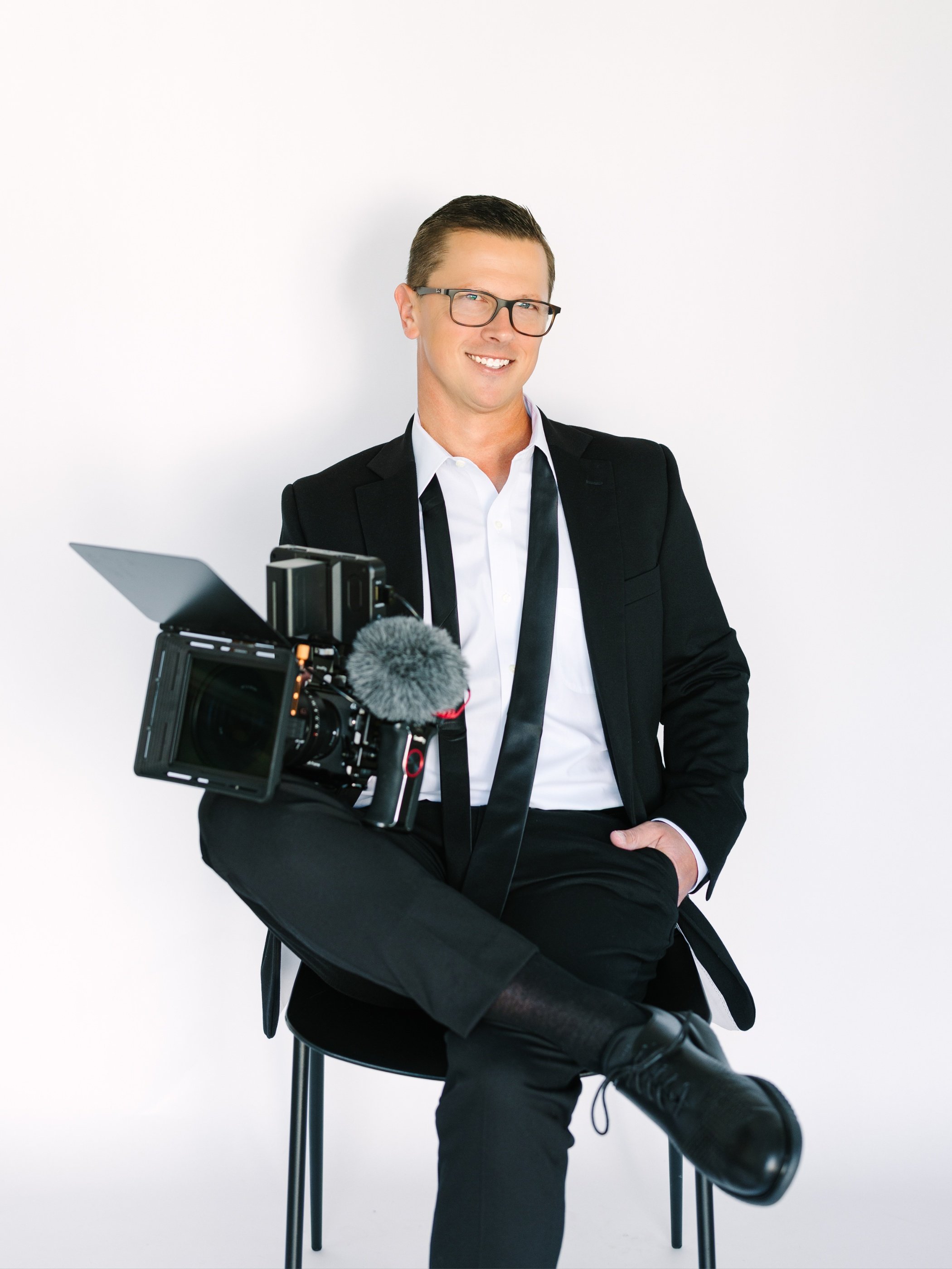 Man in a black suit with glasses sitting on a stool, with professional camera equipment hanging on his leg, smiling against a white background.