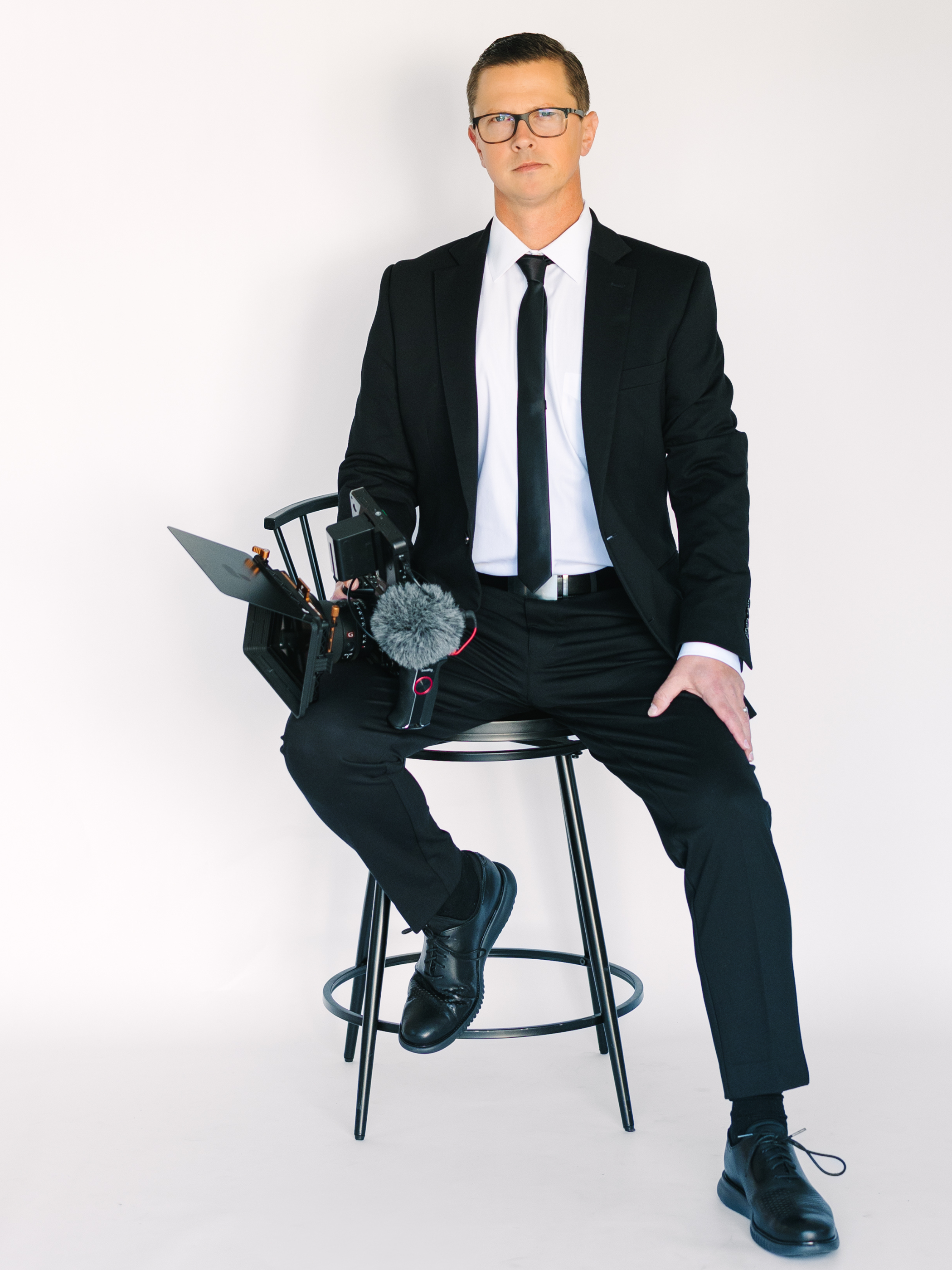 A man wearing glasses and a black suit sitting on a stool against a plain white background, holding a professional video camera with a fluffy windscreen on the microphone.