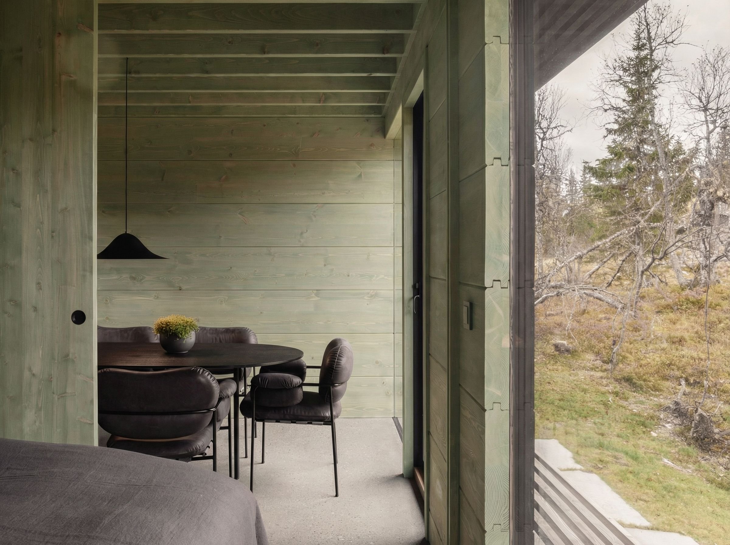 Interior of a room with a dining area featuring a round black table, five black leather chairs, a black pendant light, a small plant in a black pot on the table, and large sliding glass doors revealing a natural outdoor landscape with trees and grass.