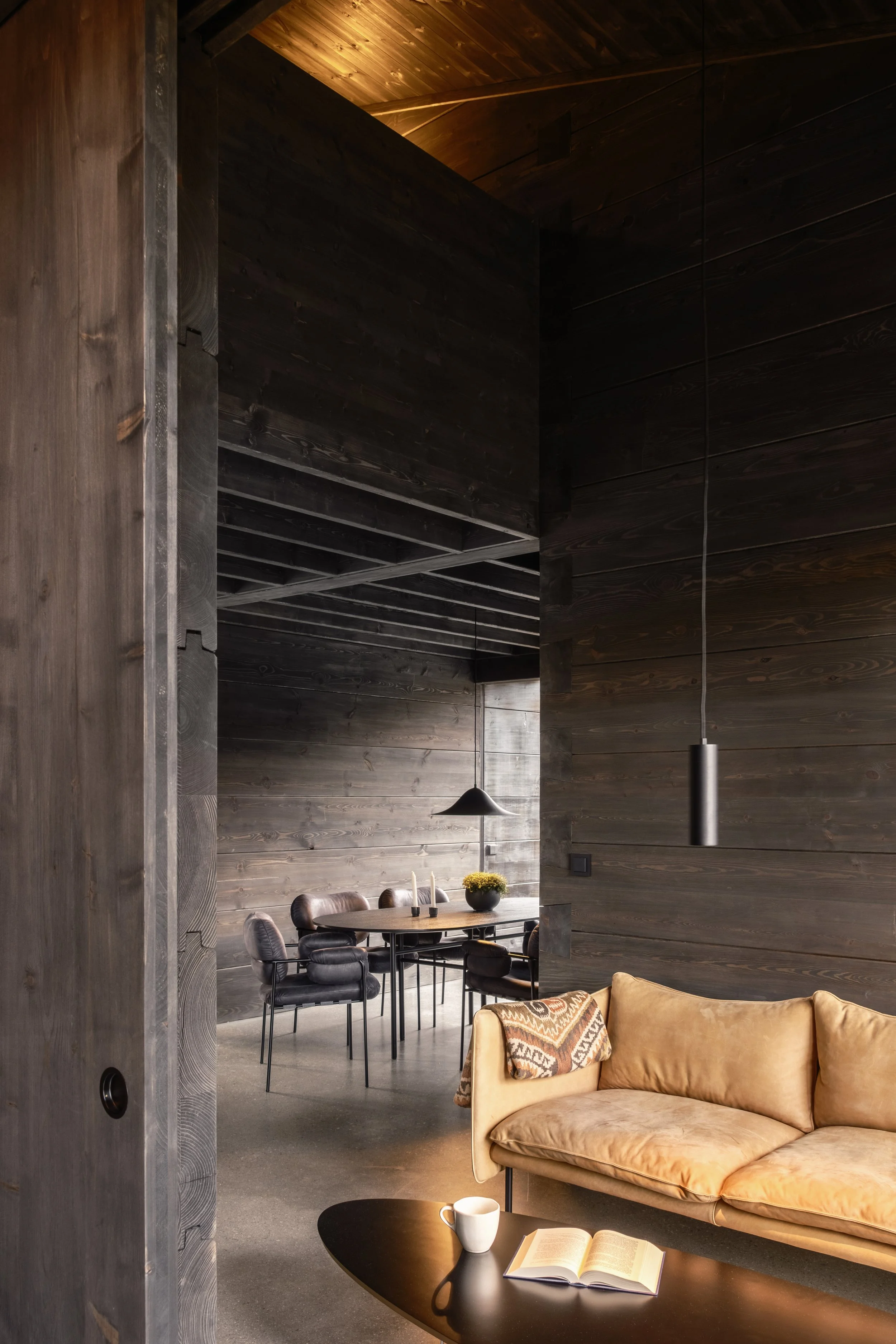 A cozy living room with a beige sofa, a black coffee table with a mug and an open book, and a dark wooden wall. In the background, a dining area with a round table, black chairs, and a black pendant light, all surrounded by dark wooden walls and ceiling.