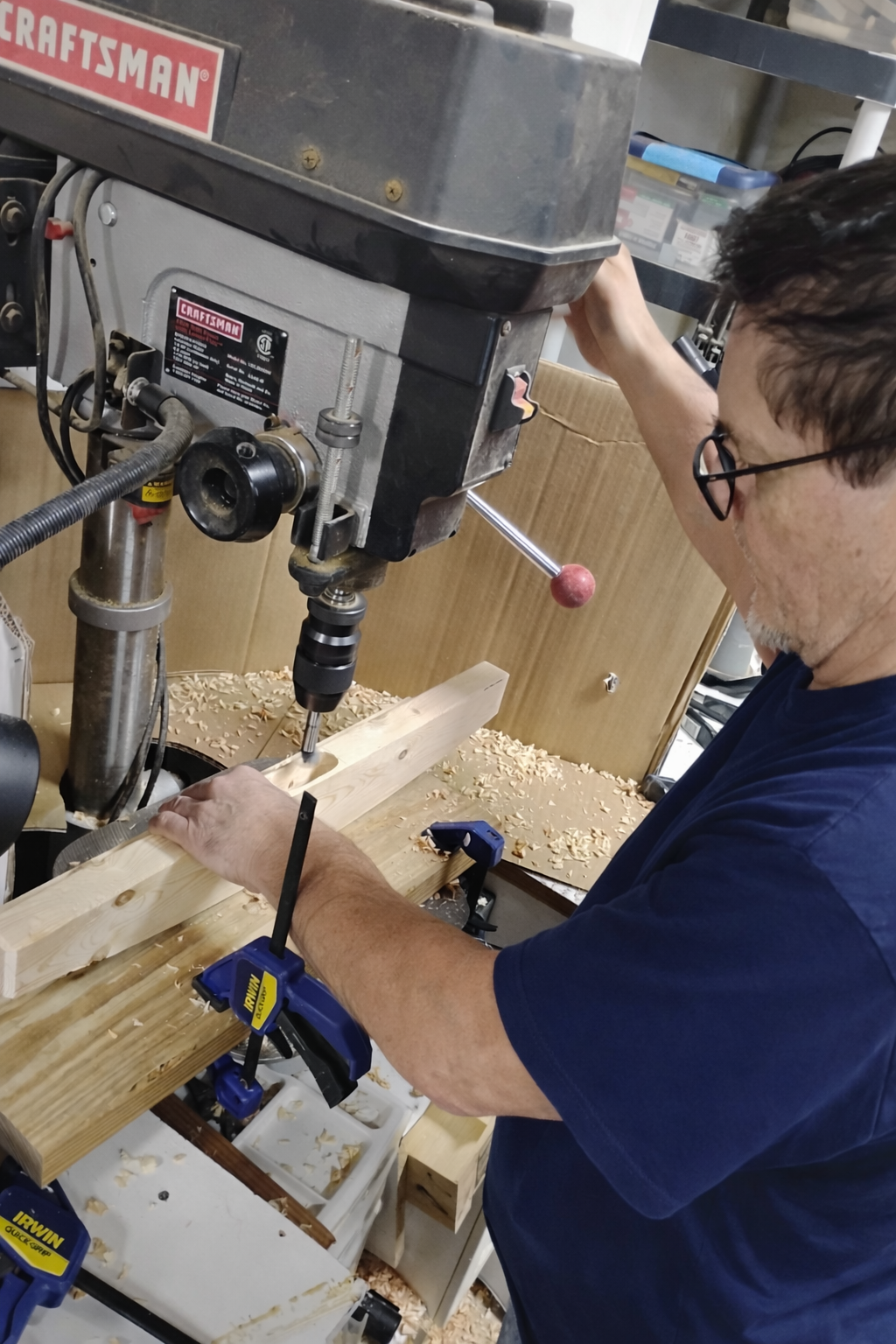 Man operating a woodworking drill press to drill into a piece of wood, with wood shavings on the work surface.