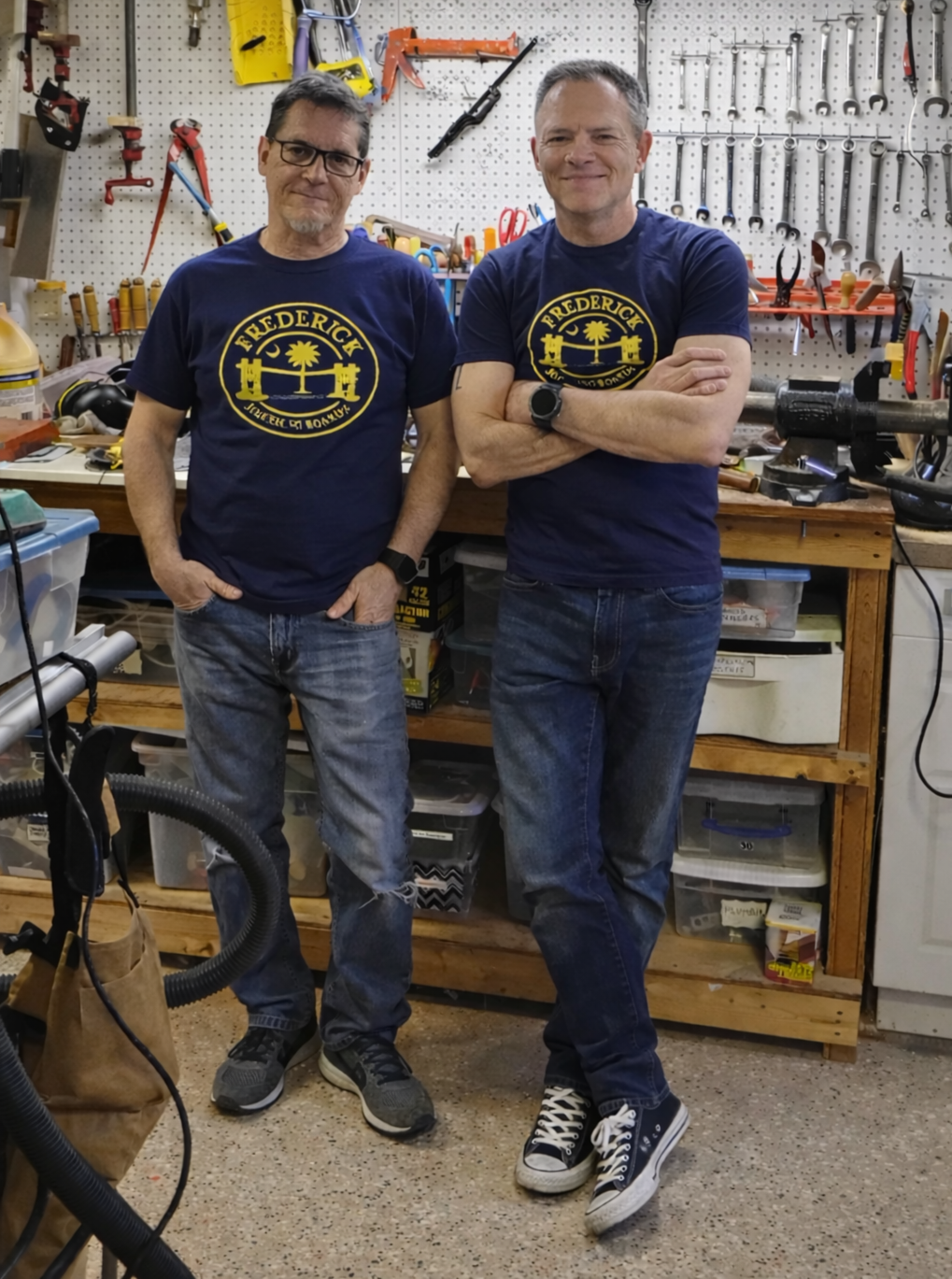 Two men standing in a workshop, both wearing navy blue t-shirts with a yellow logo featuring a palm tree and bridge, with tools and equipment on a pegboard and workbench behind them.