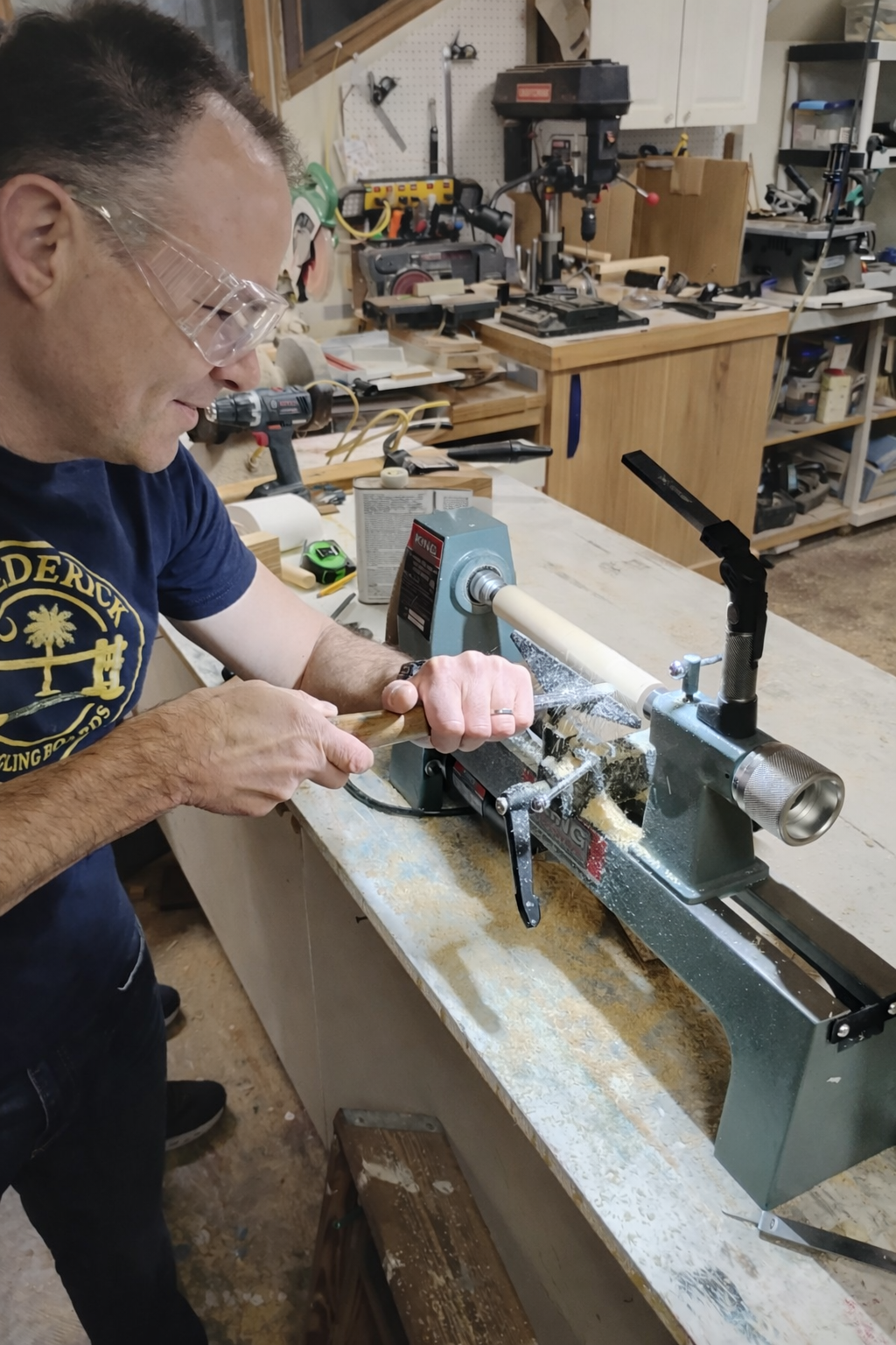 Man using a wood lathe to carve a piece of wood in a woodworking shop with various tools and equipment in the background.