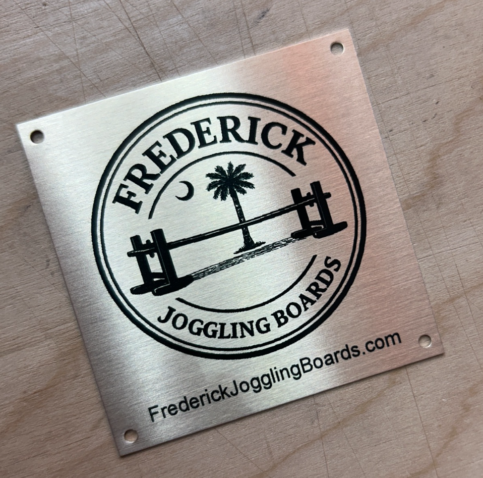 A metallic sign with the logo and website for Frederick Joggling Boards, featuring a palm tree, a crescent moon, and a joggling board.