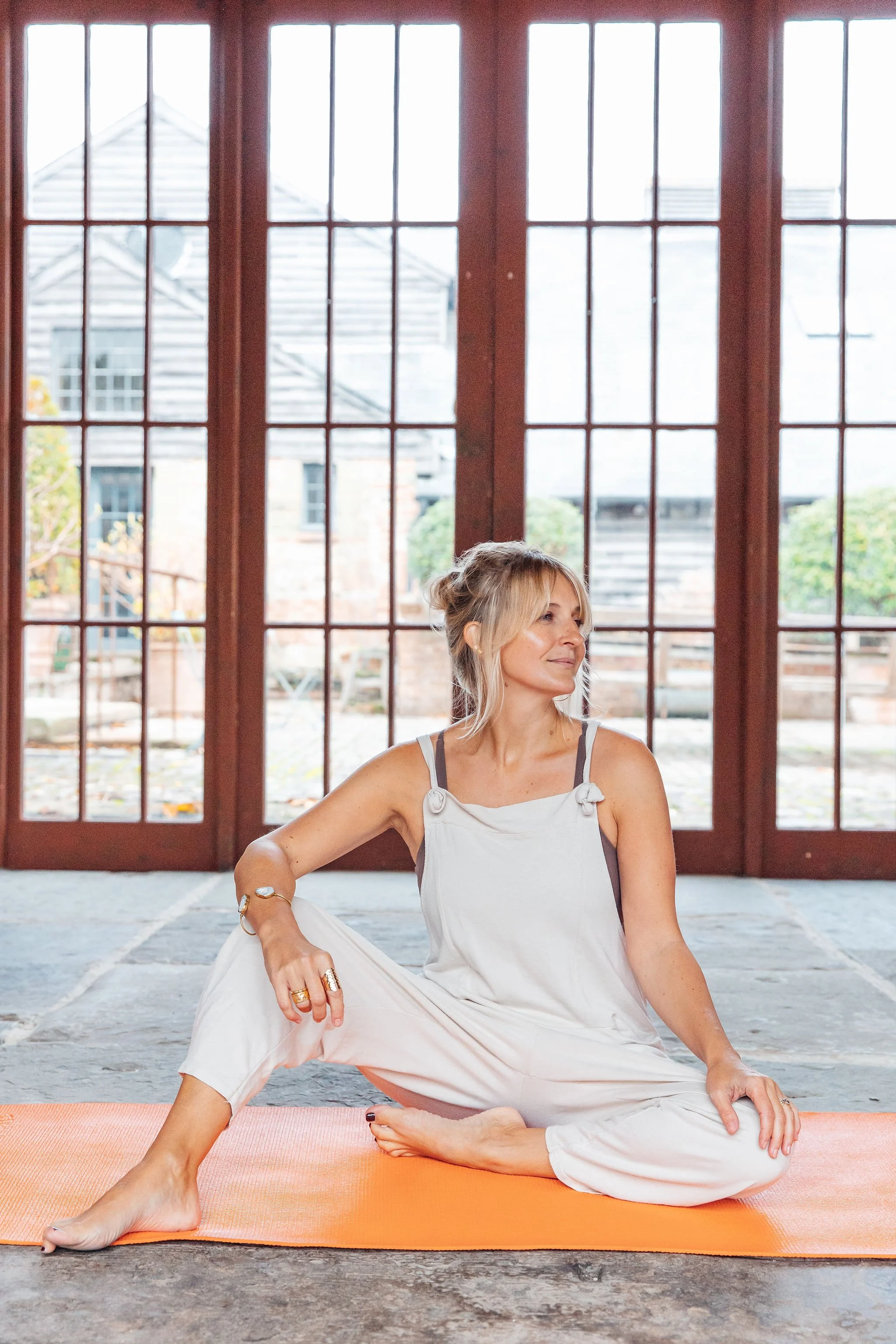 A woman practicing yoga on an orange mat inside a room with large glass doors.