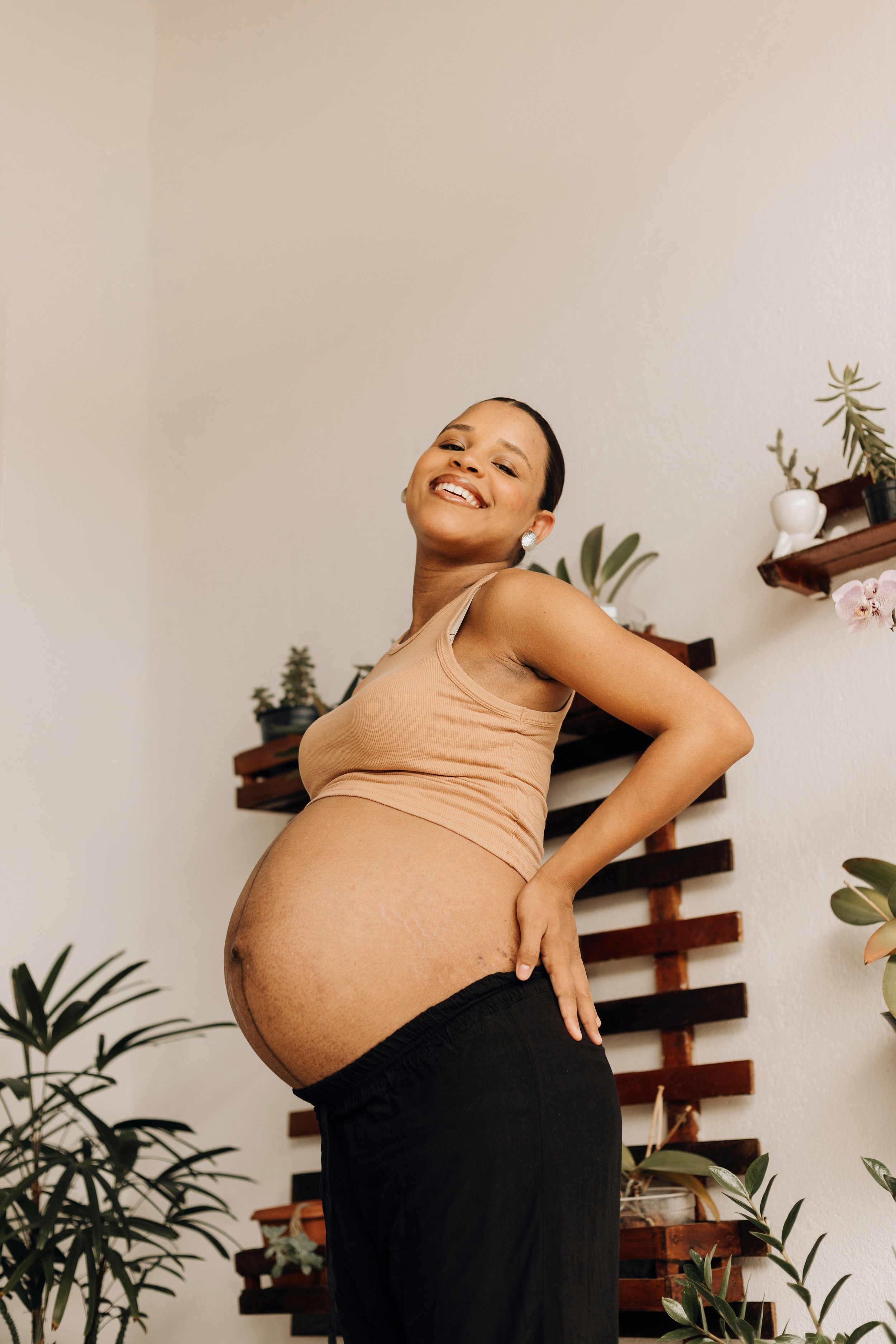 Pregnant woman smiling with hand on her baby bump indoors, with a wall and plants in the background.