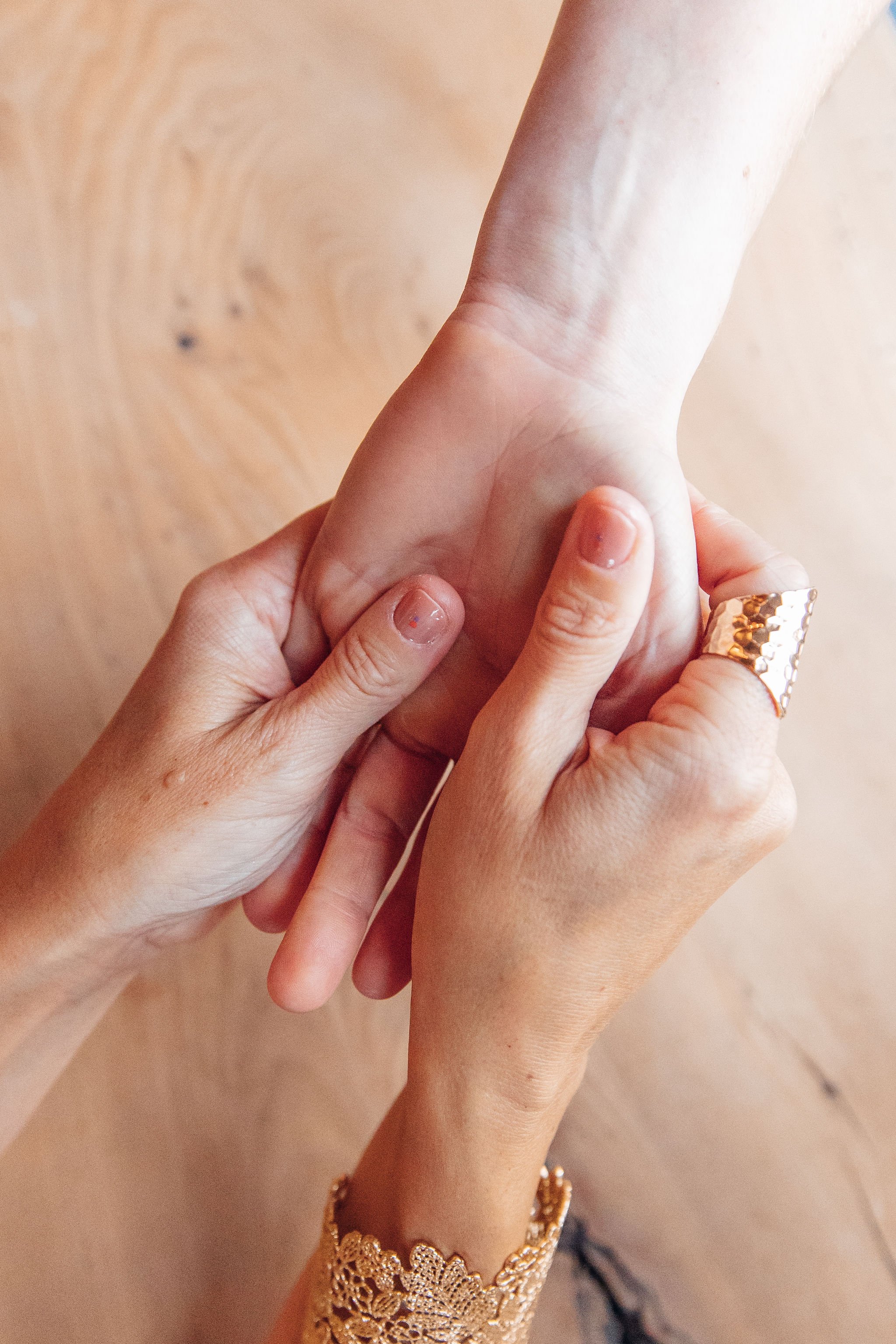 Close-up of two hands holding each other, one with a gold ring and lace sleeve, in a gentle grip over a wooden floor.