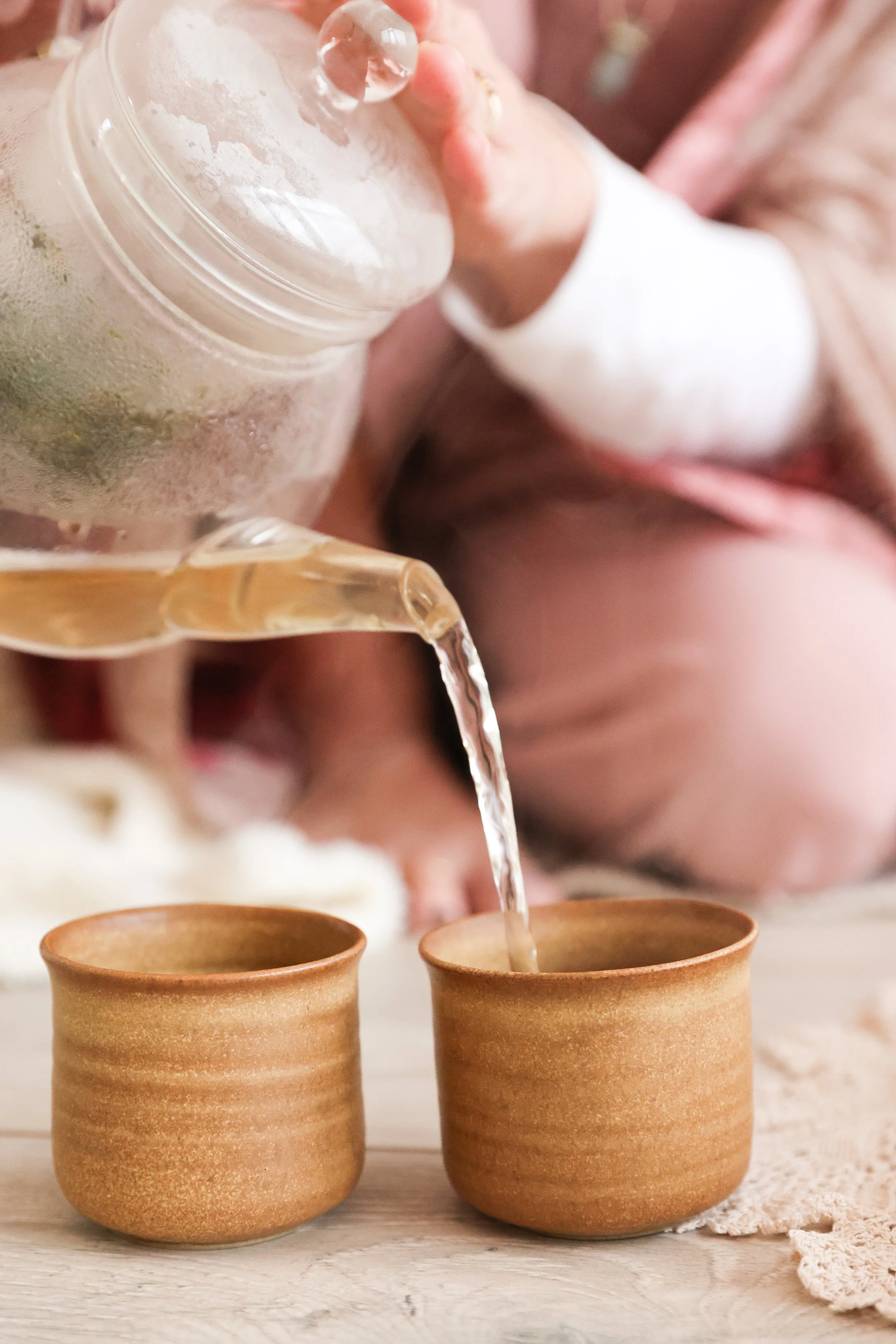 A person pouring tea from a glass teapot into two small, brown ceramic cups on a light wooden surface.
