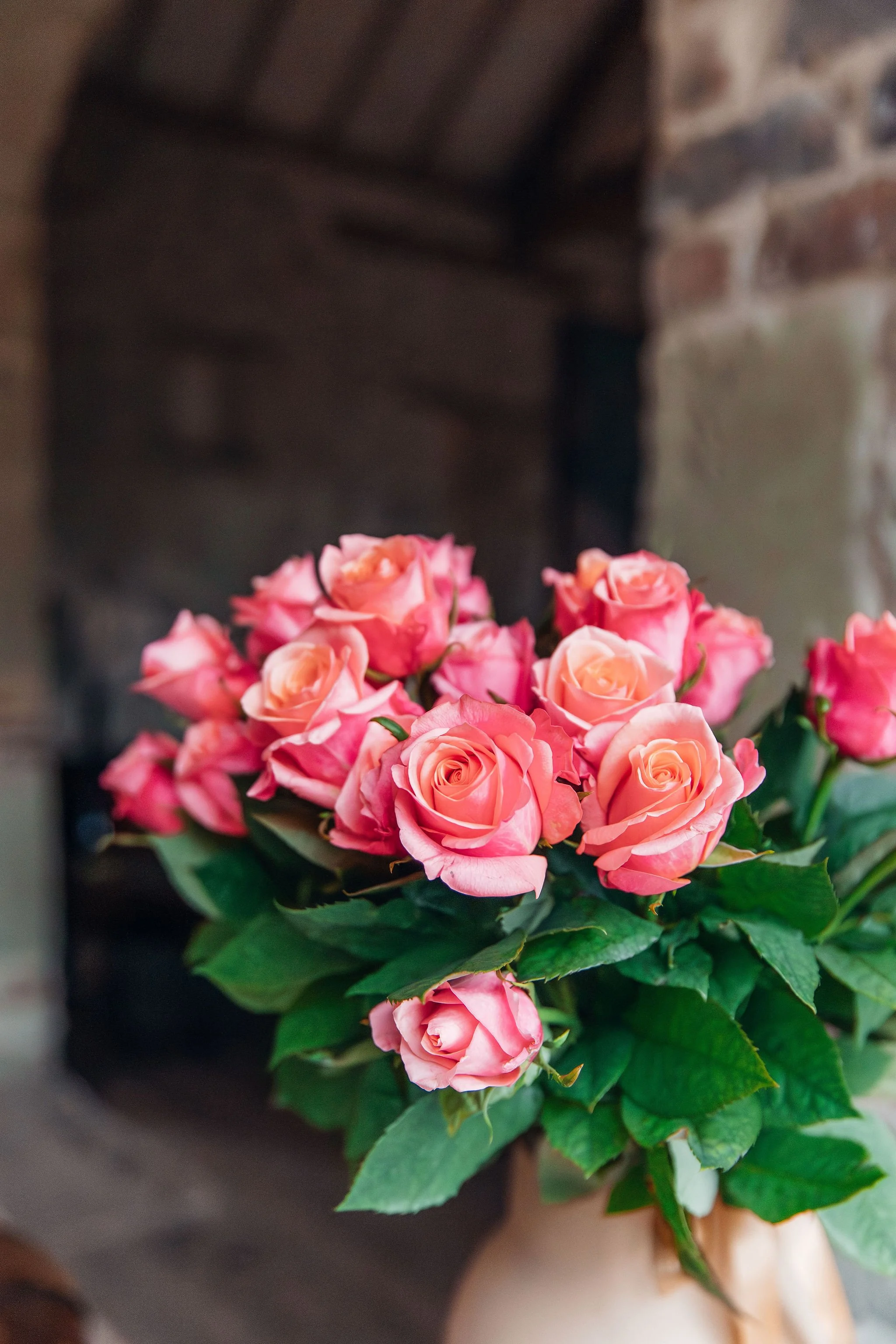 A bouquet of pink roses in a beige vase placed on a surface, with a rustic brick wall and wooden beam in the background.