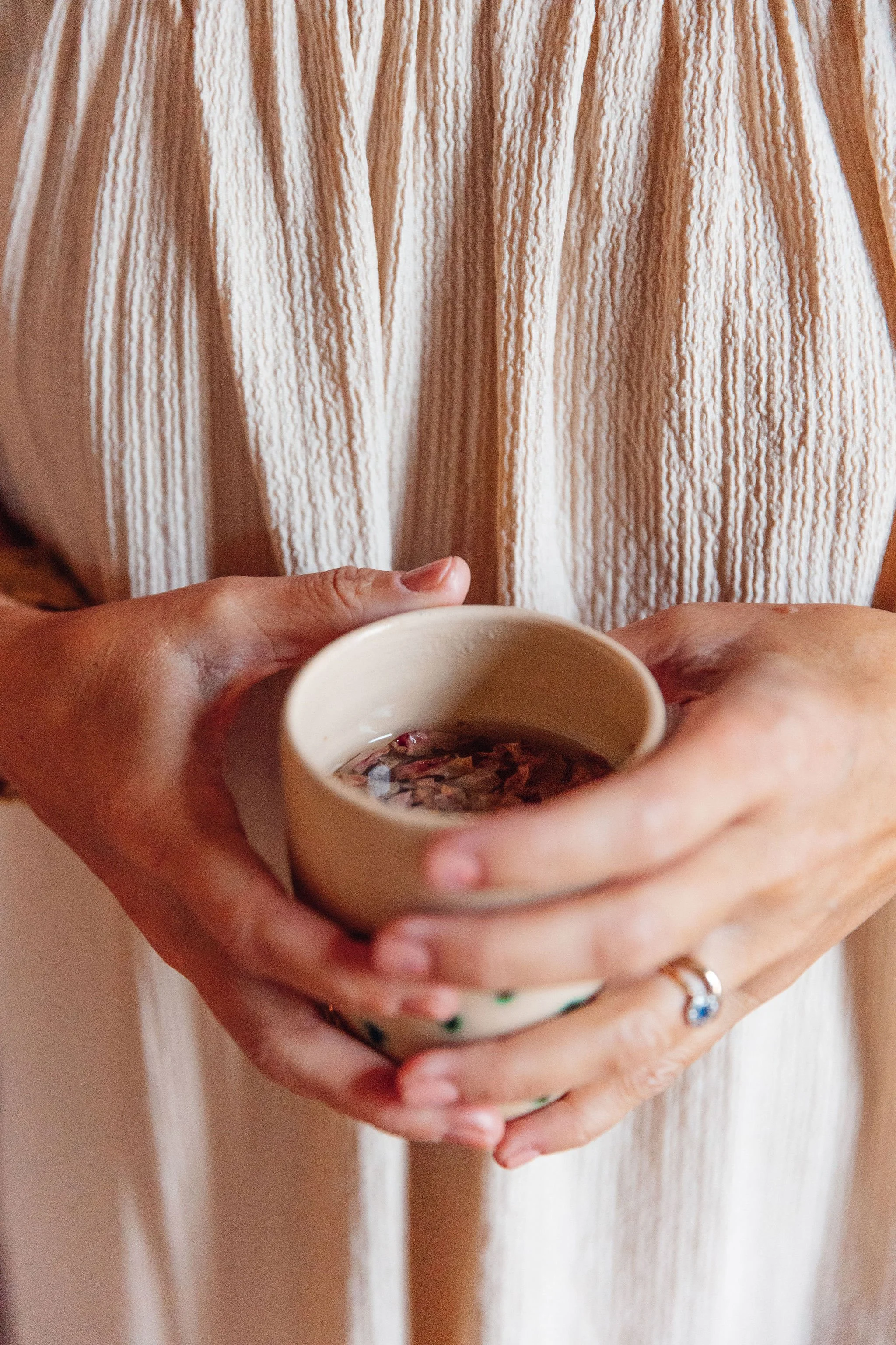 A person holding a beige mug with dried flower petals inside, wearing a ring on their finger.