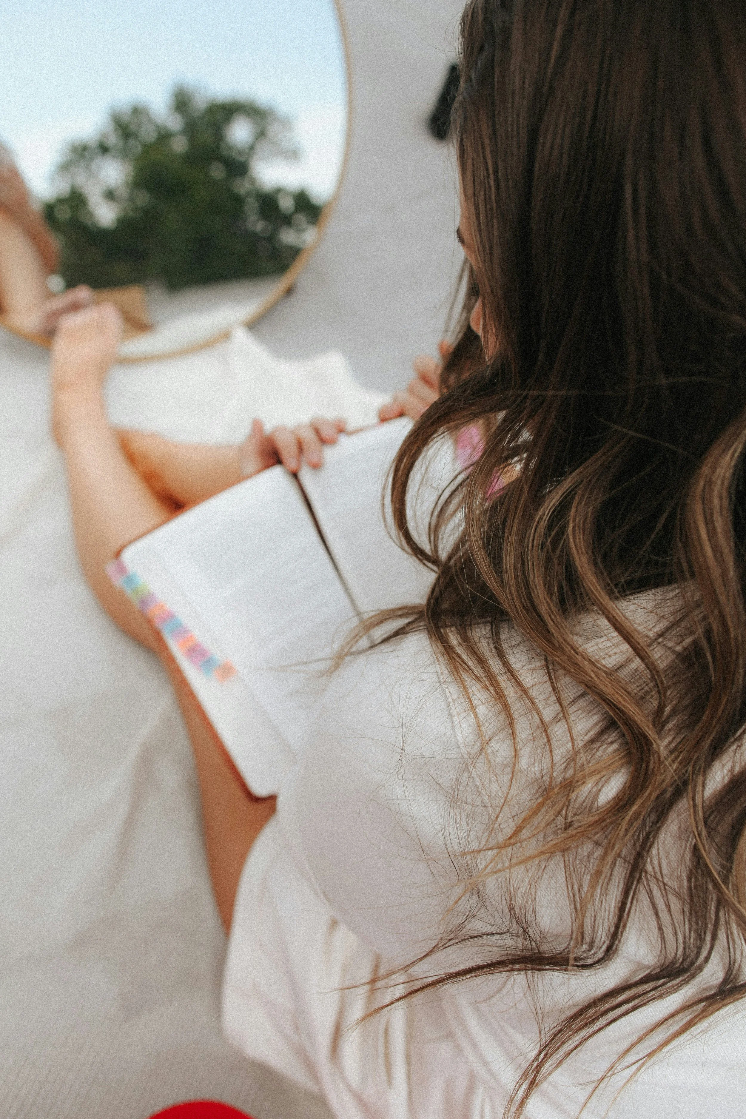 A woman with long wavy brown hair sitting on a bed, reading a book, with a mirror reflecting a tree outside the window.