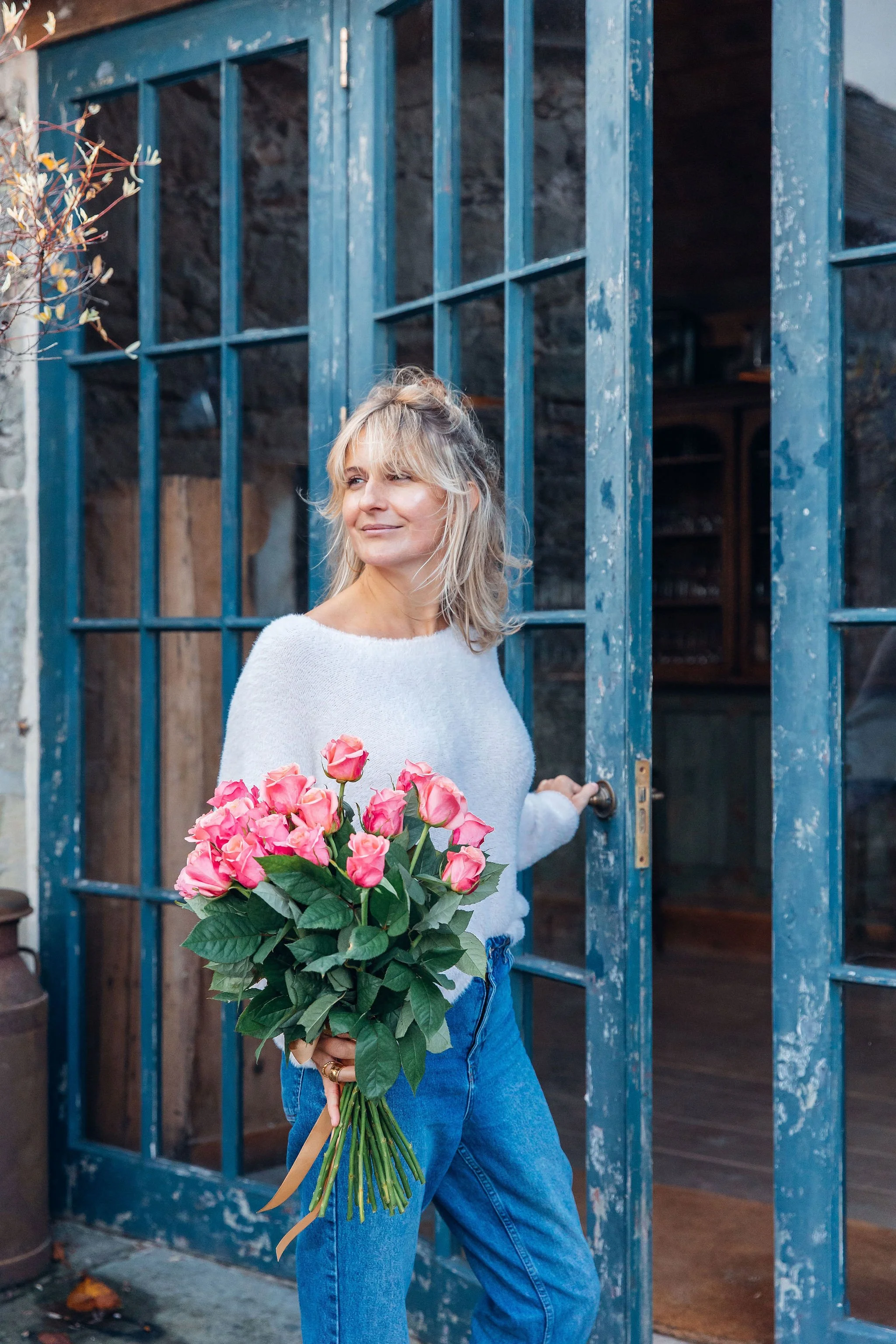 Aimee Strongman in a messy bun, wearing a white sweater and blue jeans, holding a bouquet of pink roses, standing in front of a rustic blue door.