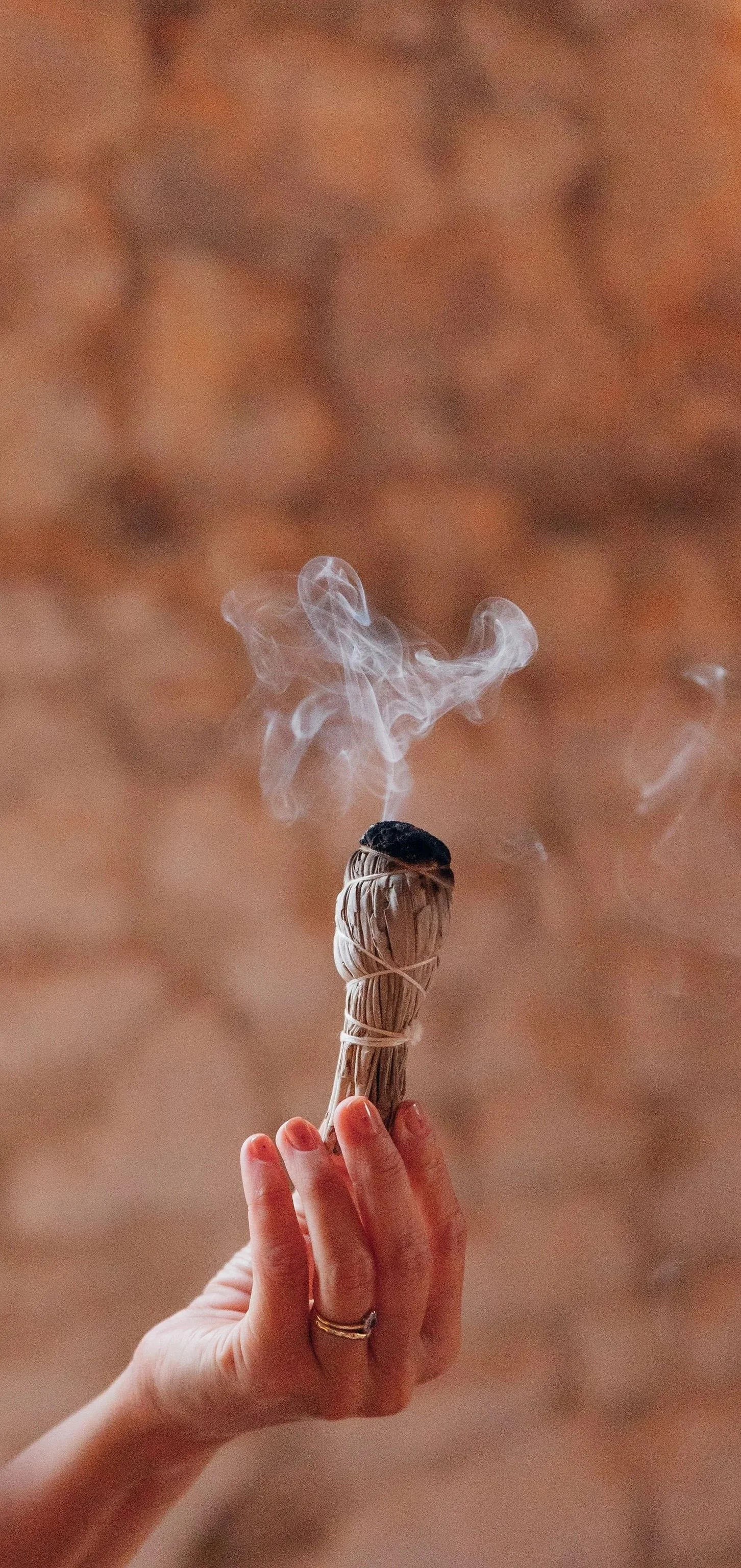 A hand holding a sage bundle with smoke rising from its burnt tip against a blurred brown background.