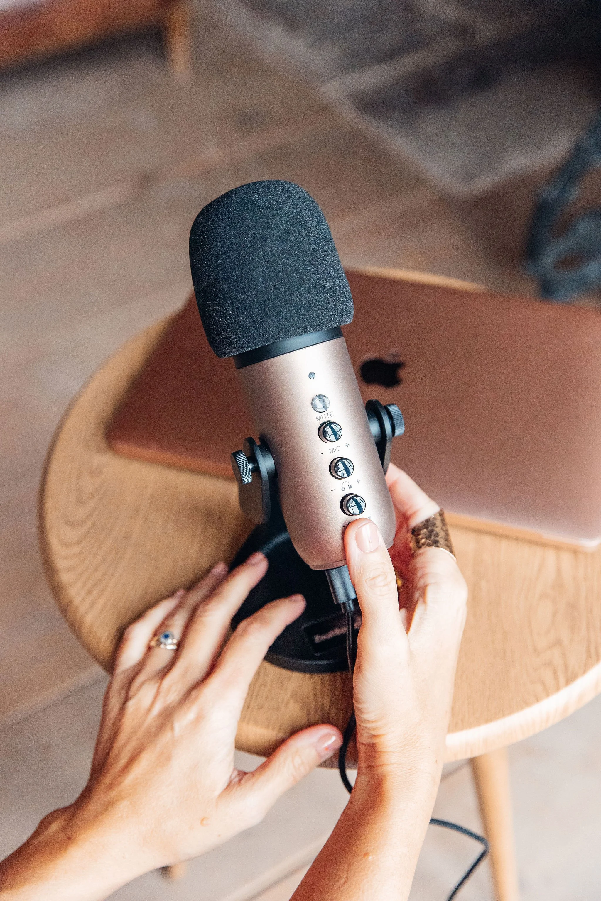 Close-up of a person's hands adjusting a silver microphone on a wooden table, with a pink laptop in the background.