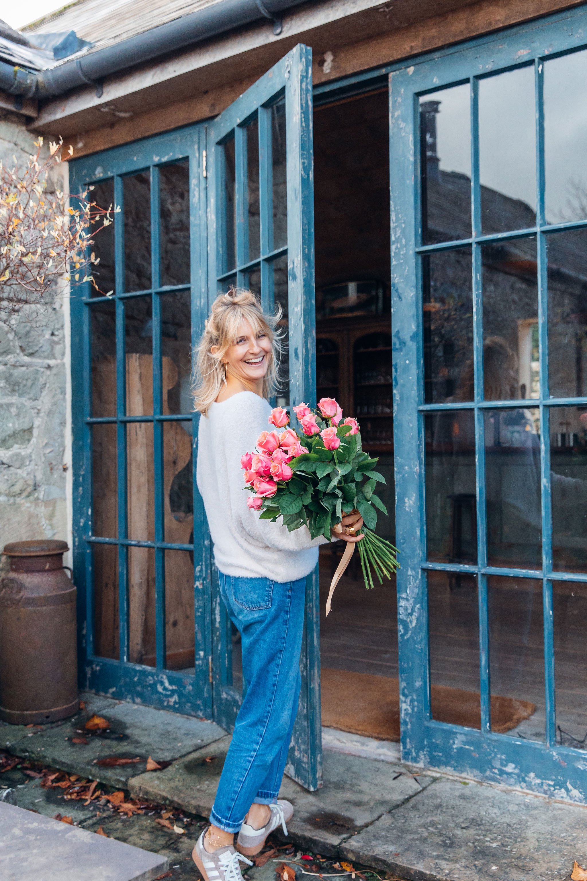 A woman standing outside a rustic building holding a bouquet of pink roses, smiling and looking back.