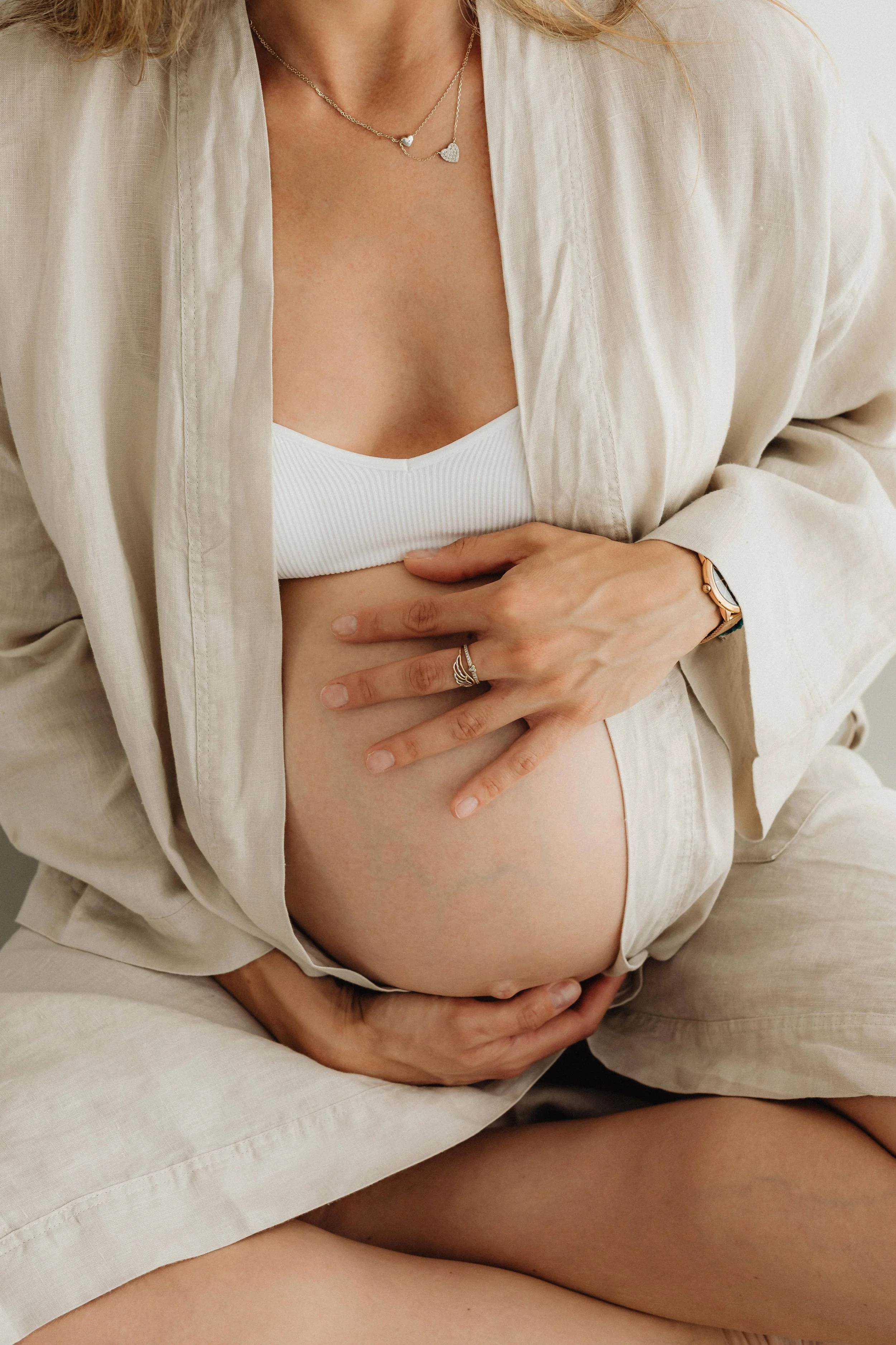 A pregnant woman wearing a white tank top and beige open shirt, sitting and holding her belly with both hands. She has jewelry including a ring, a watch, and a necklace, and is in a neutral-colored setting.