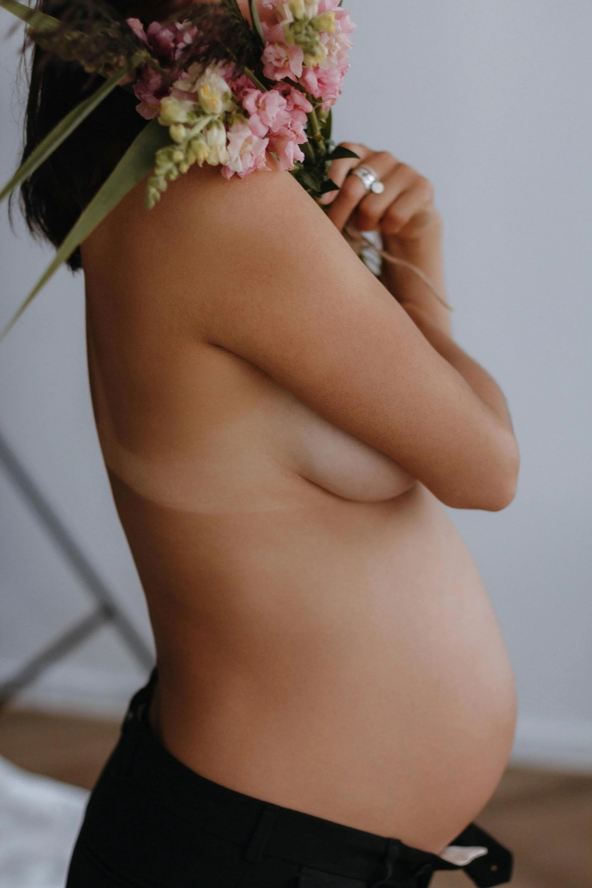 A pregnant woman holding a bouquet of pink flowers to her shoulder, with her arm crossed in front of her chest, wearing black shorts.