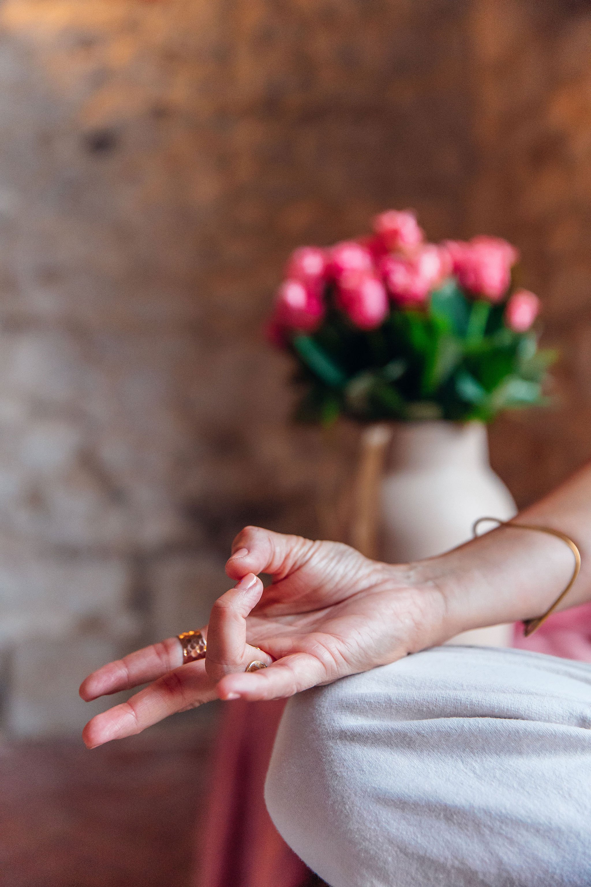A person's hand resting on their lap with a blurred bouquet of pink flowers in a ceramic vase in the background.