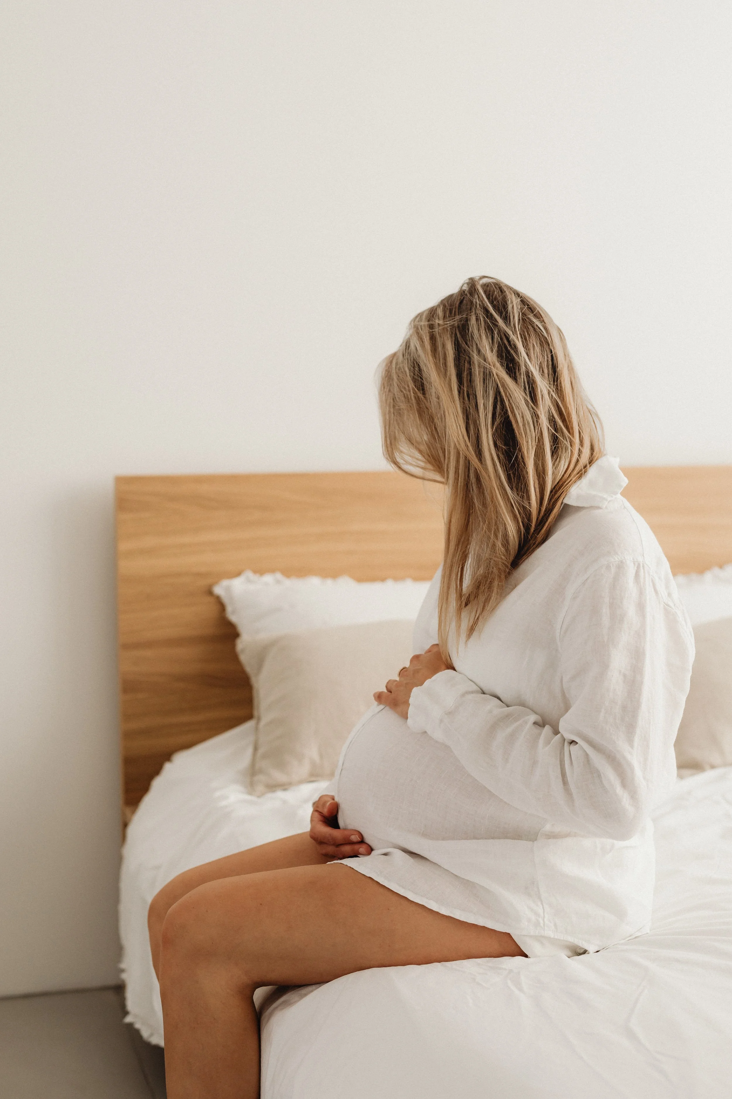A pregnant woman sitting on a bed, cradling her belly with both hands, in a minimalist bedroom with white walls and wooden headboard.
