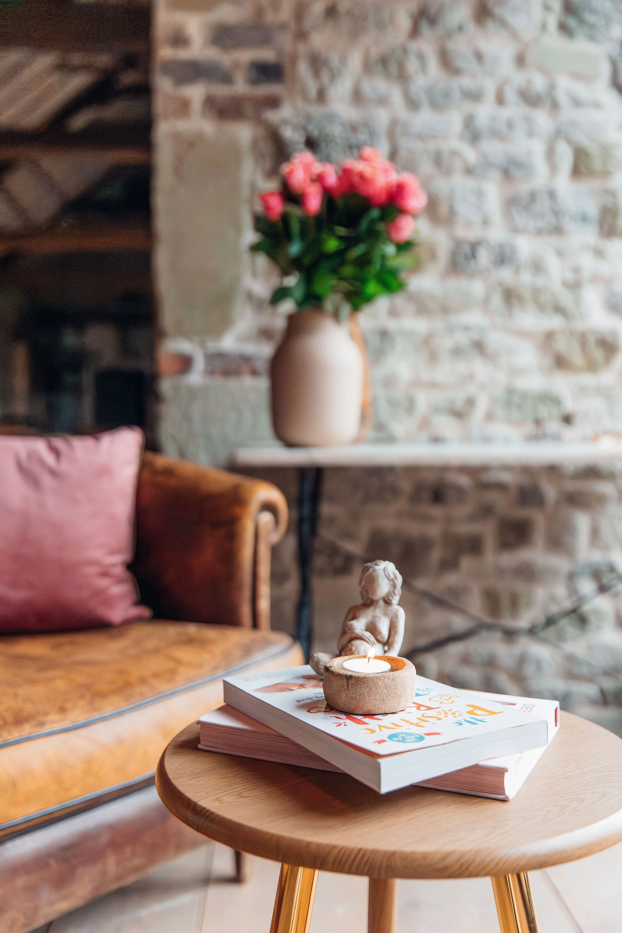A cozy living room scene with a wooden side table holding a lit candle, a small sculpture, and a stack of magazines, with a leather couch and a vase of pink flowers in the background.