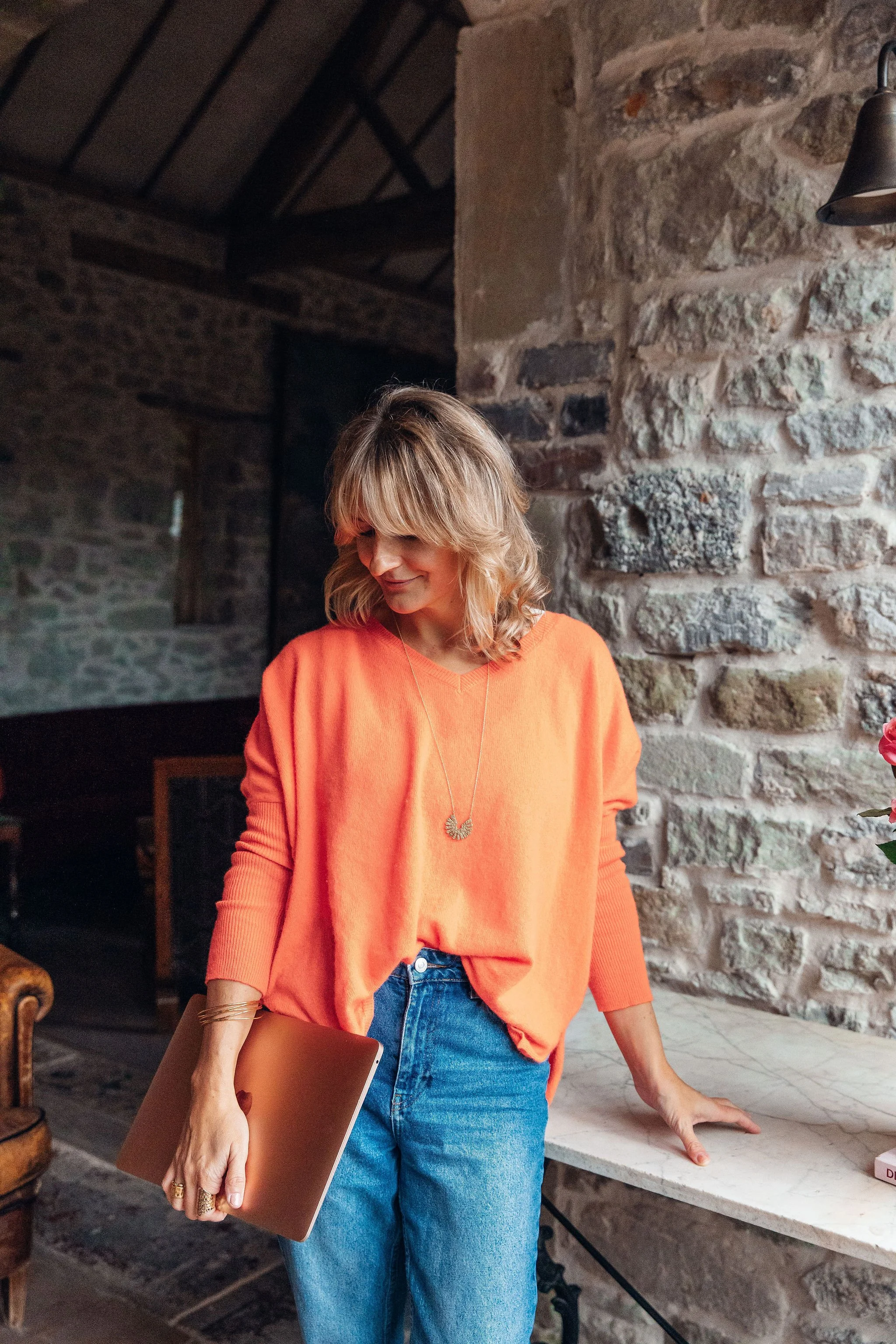 Aimee Strongman in an orange sweatshirt and jeans holding a laptop stands in a rustic room with exposed stone walls.