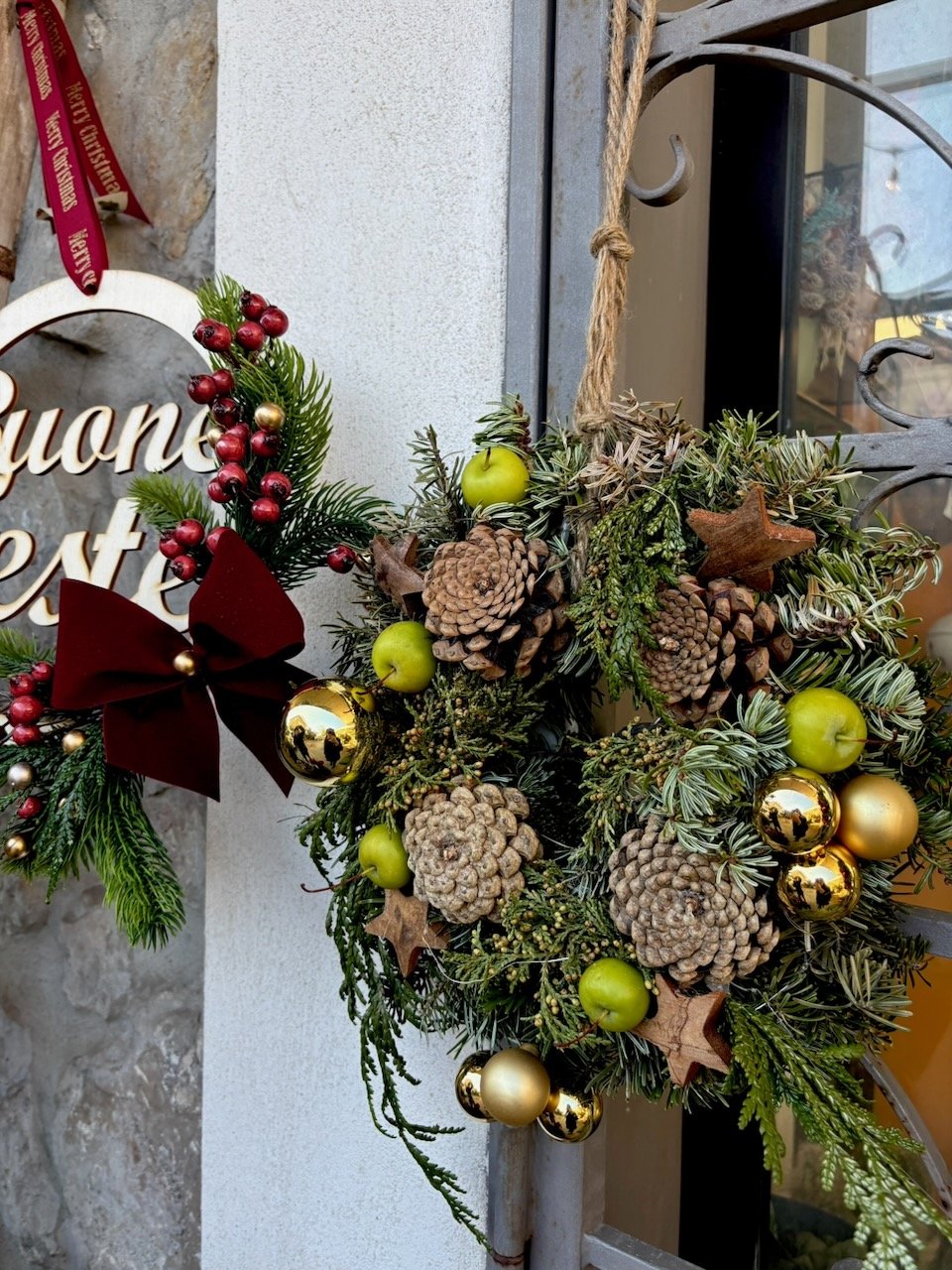 Coroncina natalizia decorata con pigne, mele verdi, bacche rosse, stelle di legno, palline dorate e verdi, fiocco di velluto rosso, appesa a una porta con un cordino di spago.