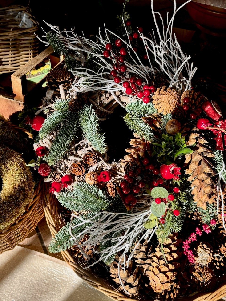 Corona natalizia decorata con pigne, rami di pino, bacche rosse e foglie verdi, ambientata in un cestino di vimini.