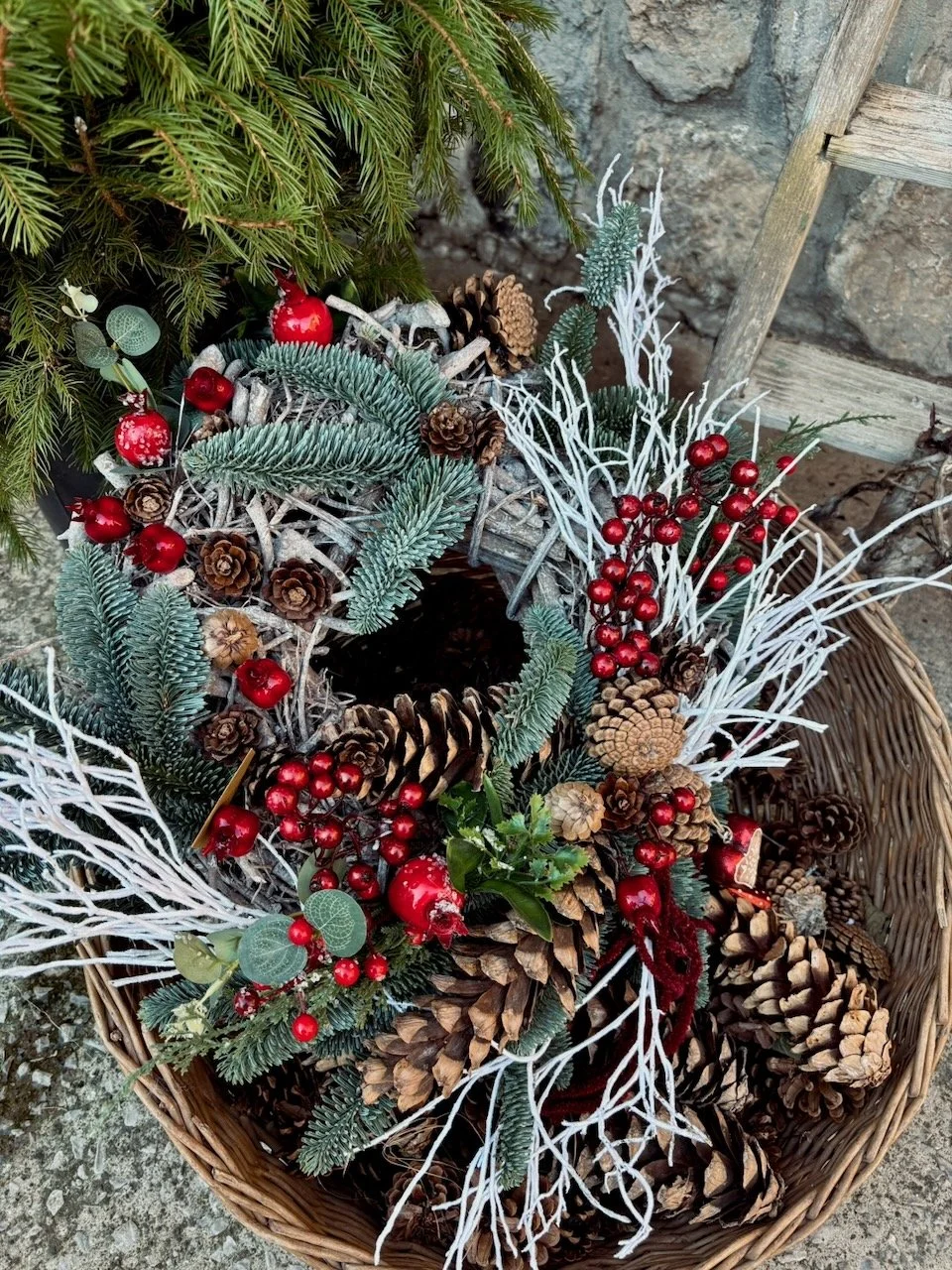 Corona decorativa natalizia con pigne, rami di abete, bacche rosse, rattan e decorazioni bianche.