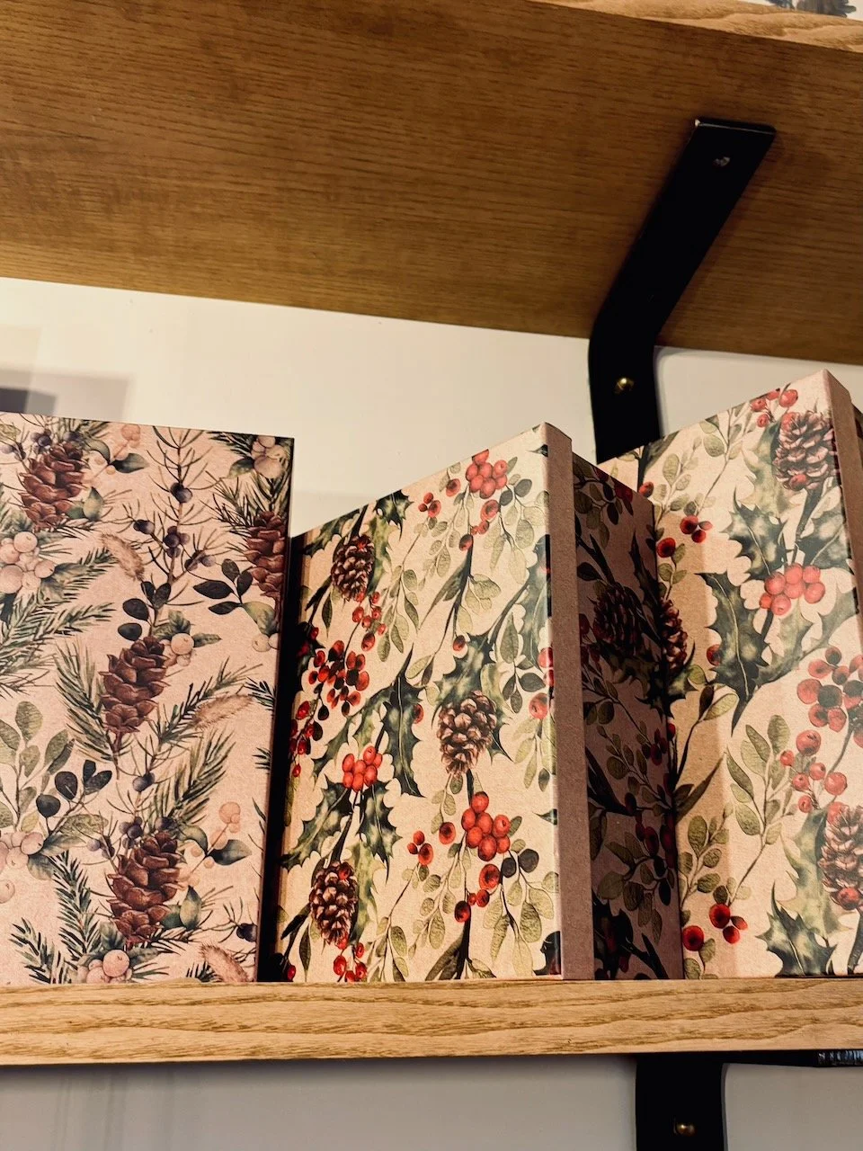 Scatole decorate con motivi natalizi di pigne, bacche rosse e foglie di agrifoglio su uno scaffale di legno.