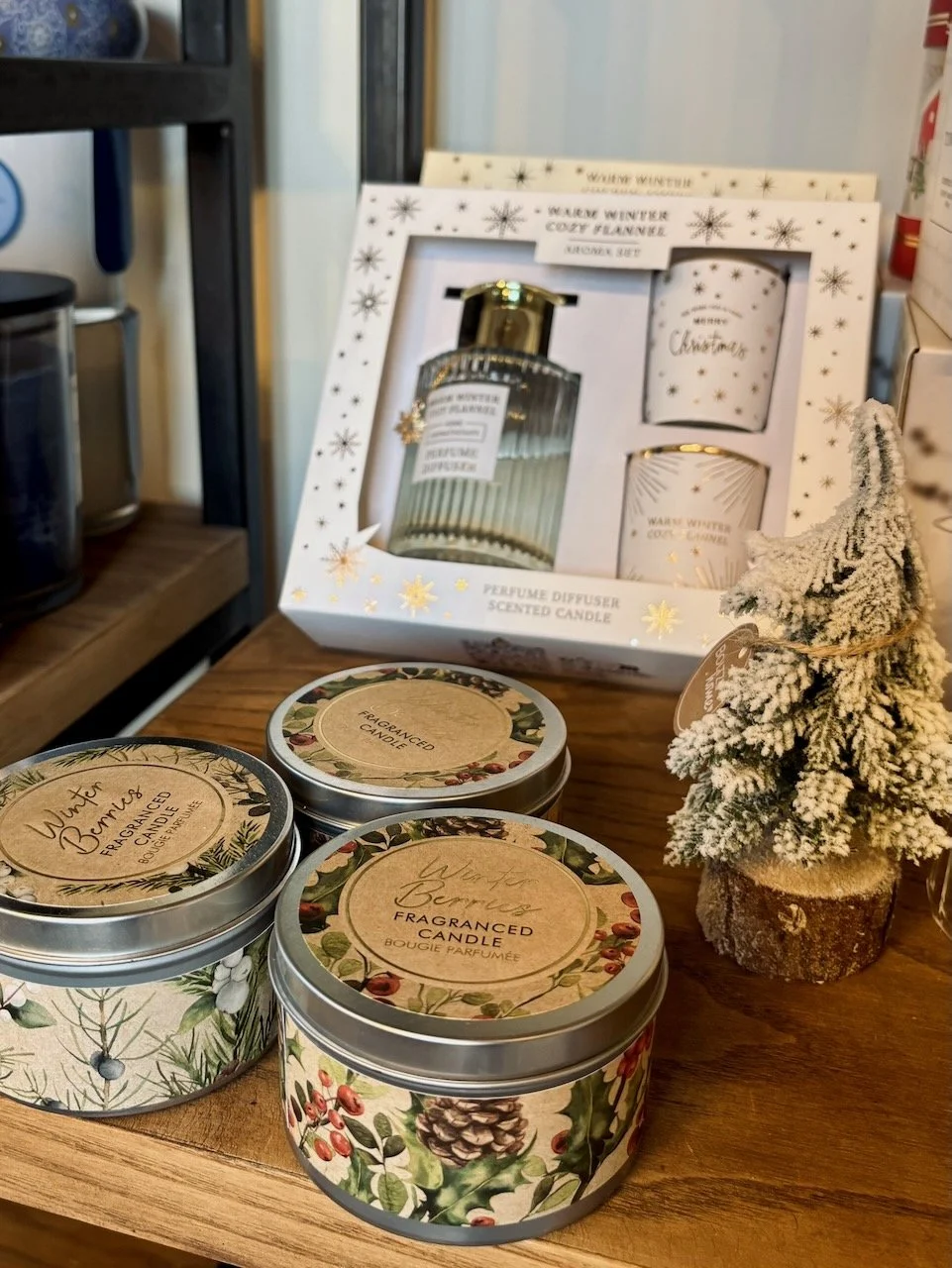 Tre scatole di candele profumate con decorazioni natalizie, un piccolo albero di Natale innevato e un set di diffusore di profumo natalizio confezionato, su un tavolo di legno.