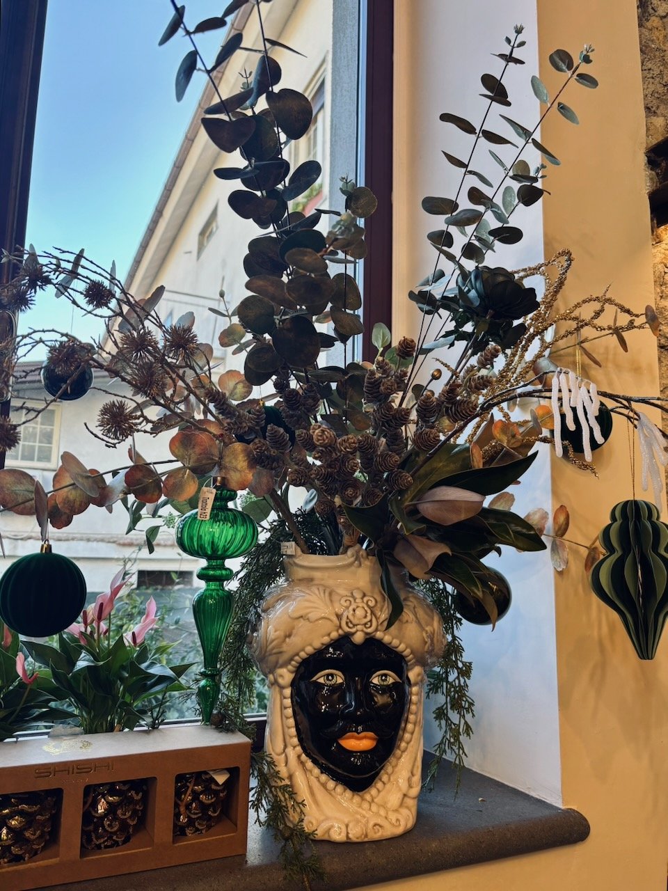 Un vaso decorativo molto particolare con una faccia nera e dettagli color carne, riempito di vari rami e foglie verdi, posizionato vicino a una finestra illuminate dalla luce naturale.