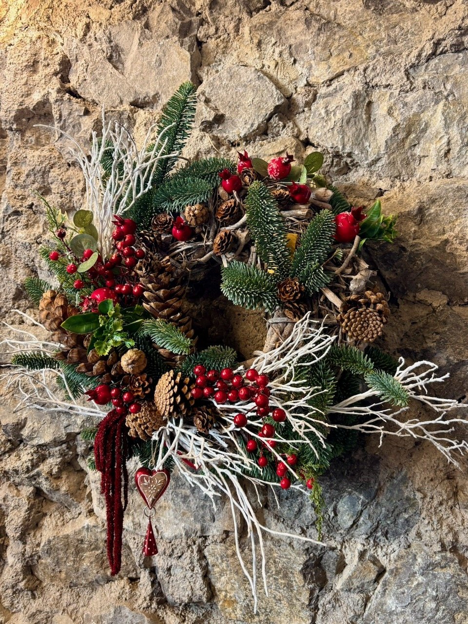 Corona natalizia decorata con pigne, rami di abete, bacche rosse, ramoscelli bianchi, foglie verdi, e un cuore decorativo, appesa a un muro di pietra.