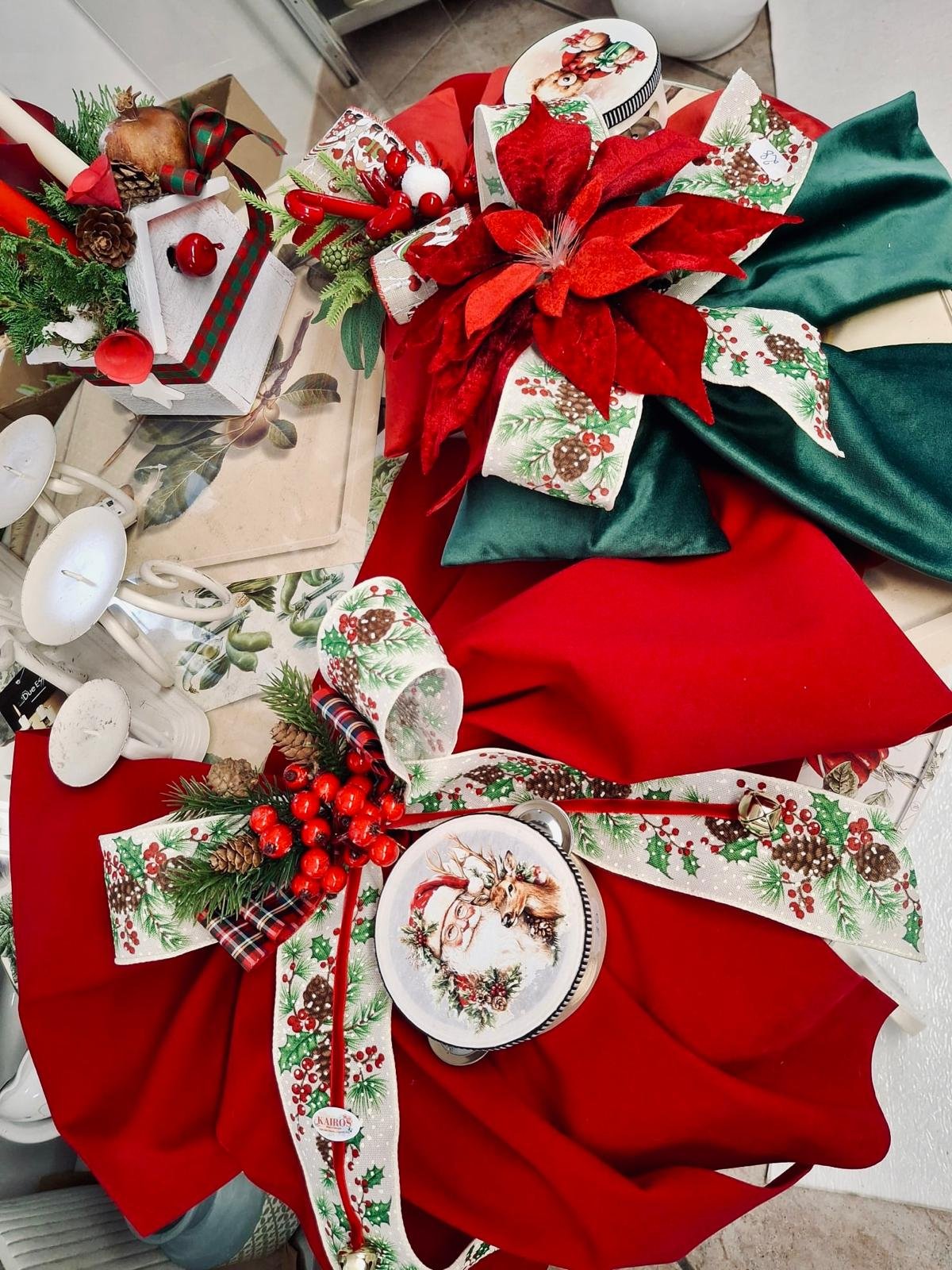 Decorazioni natalizie su un tavolo, tra cui un vaso con piante verdi e bacche rosse, un piatto con disegno di Rudolph il renna, fiocchi decorativi con motivi natalizi, e tovaglie rosse e verdi