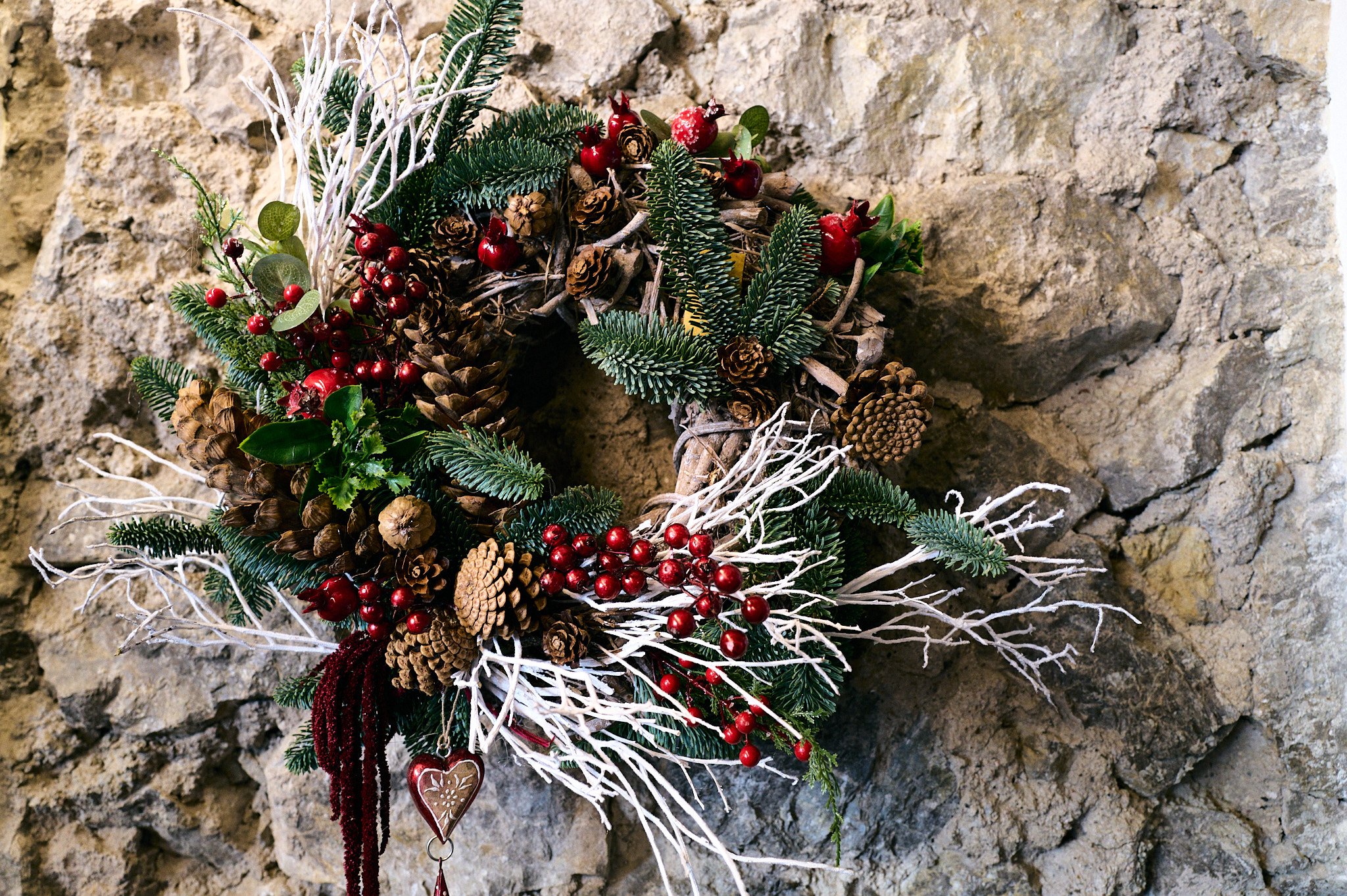 Corona di Natale decorata con pigne, rami di abete, bacche rosse e elementi naturali appesa a un muro di pietra.