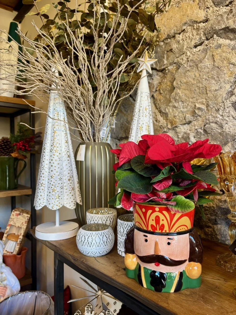 Decorazioni natalizie, tra cui un vaso con rami secchi decorativi, un orsetto contenitore portapiante con un pino natalizio rosso, e candele decorative con motivi geometrici, tutto disposto su un tavolo in un ambiente interno con parete di pietra.